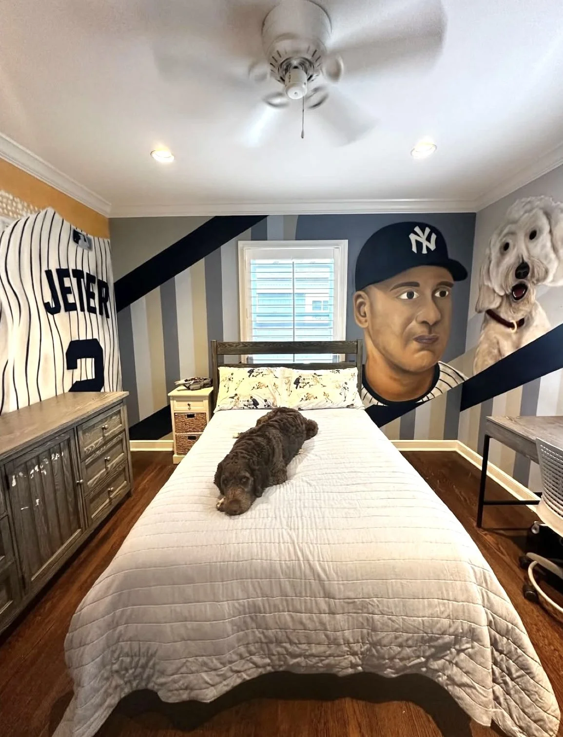Bedroom with baseball-themed decor, featuring wall art of a dog and a baseball player, a dog resting on the bed, and furniture including a wooden dresser, nightstand, and a desk with a chair.