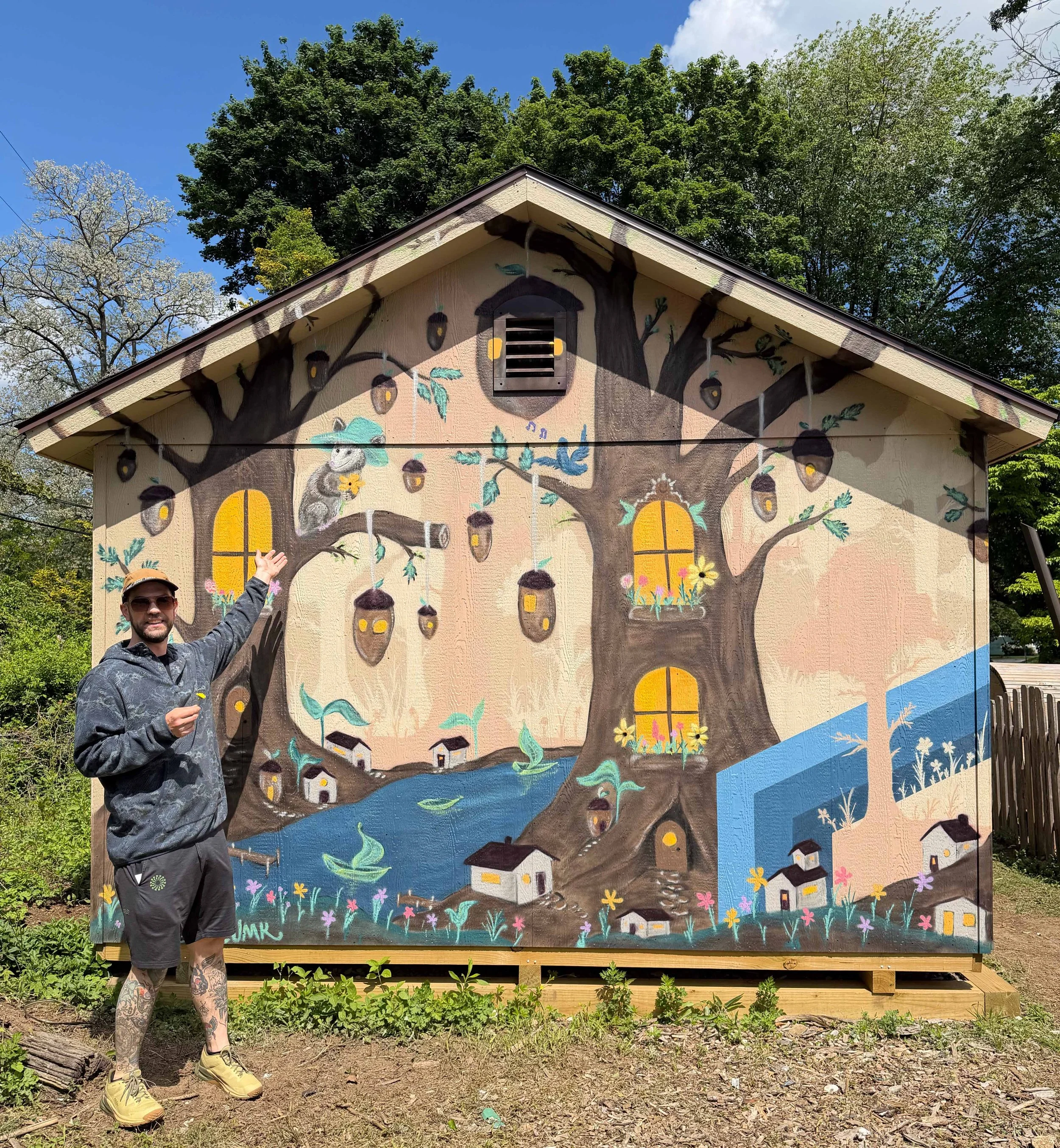 Man with tattoos pointing at a mural depicting a large tree with houses and animals painted on outdoor building wall