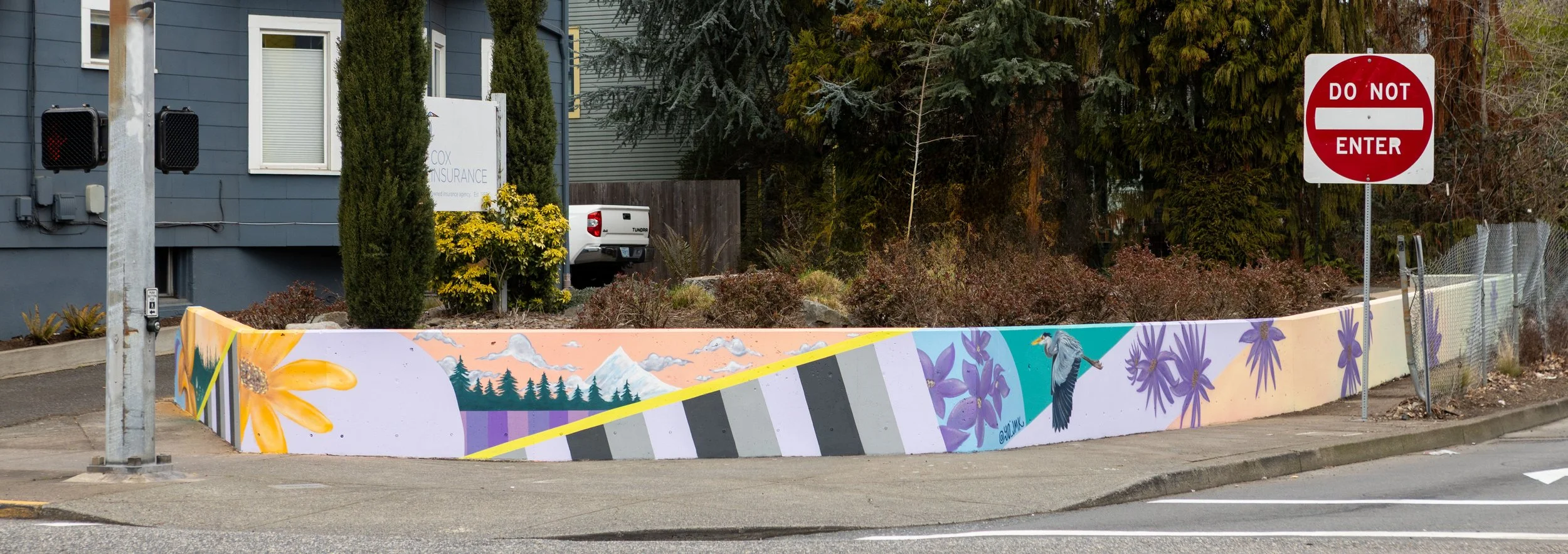 Colorful mural on a low wall featuring flowers, mountains, trees, a bird, and a sky with clouds at street corner with a 'Do Not Enter' sign.