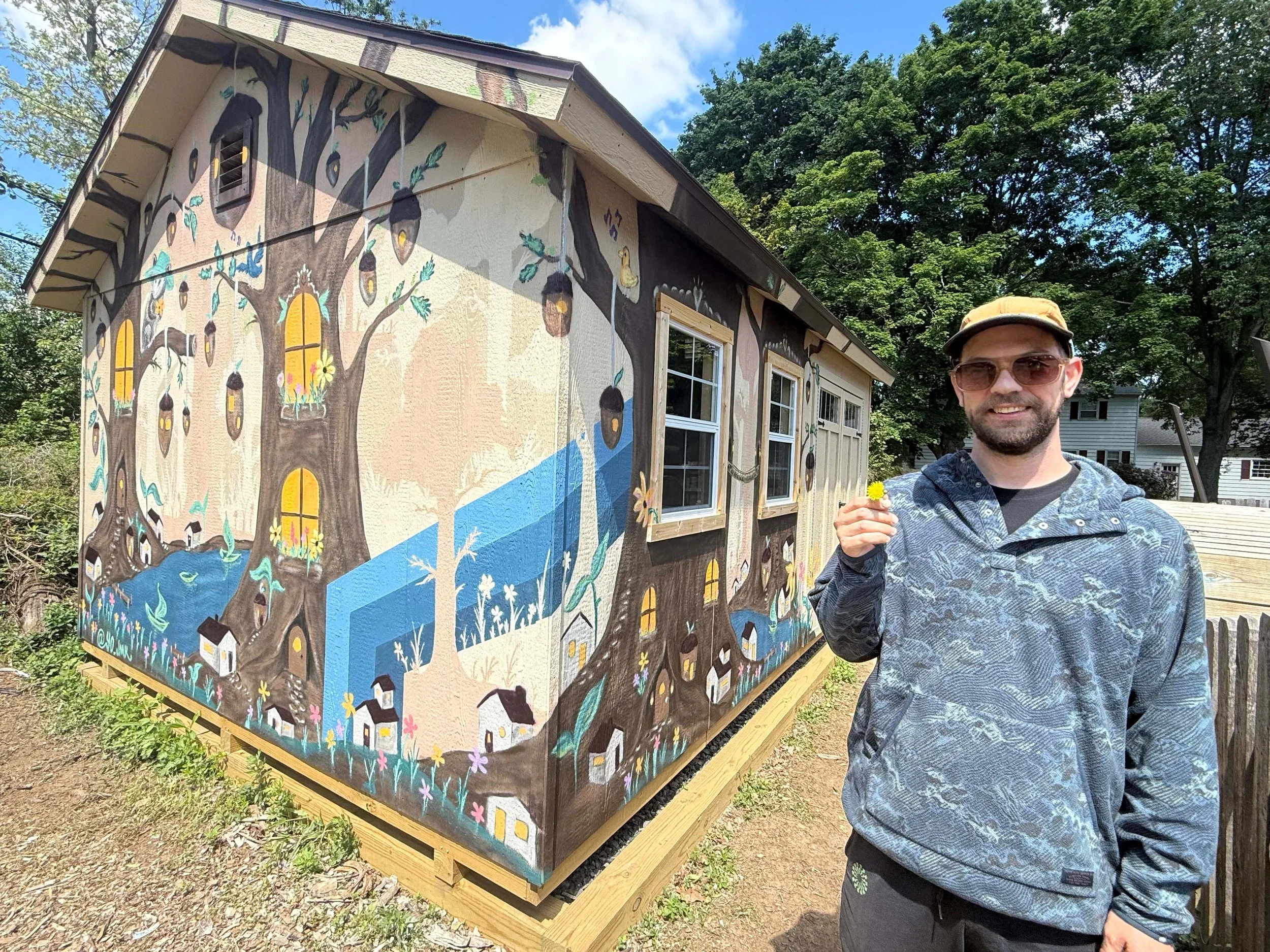 A man wearing sunglasses and a cap holding a yellow dandelion flower standing in front of a house with a mural painted on the exterior wall depicting a large tree with lanterns hanging from its branches and a small village underneath.