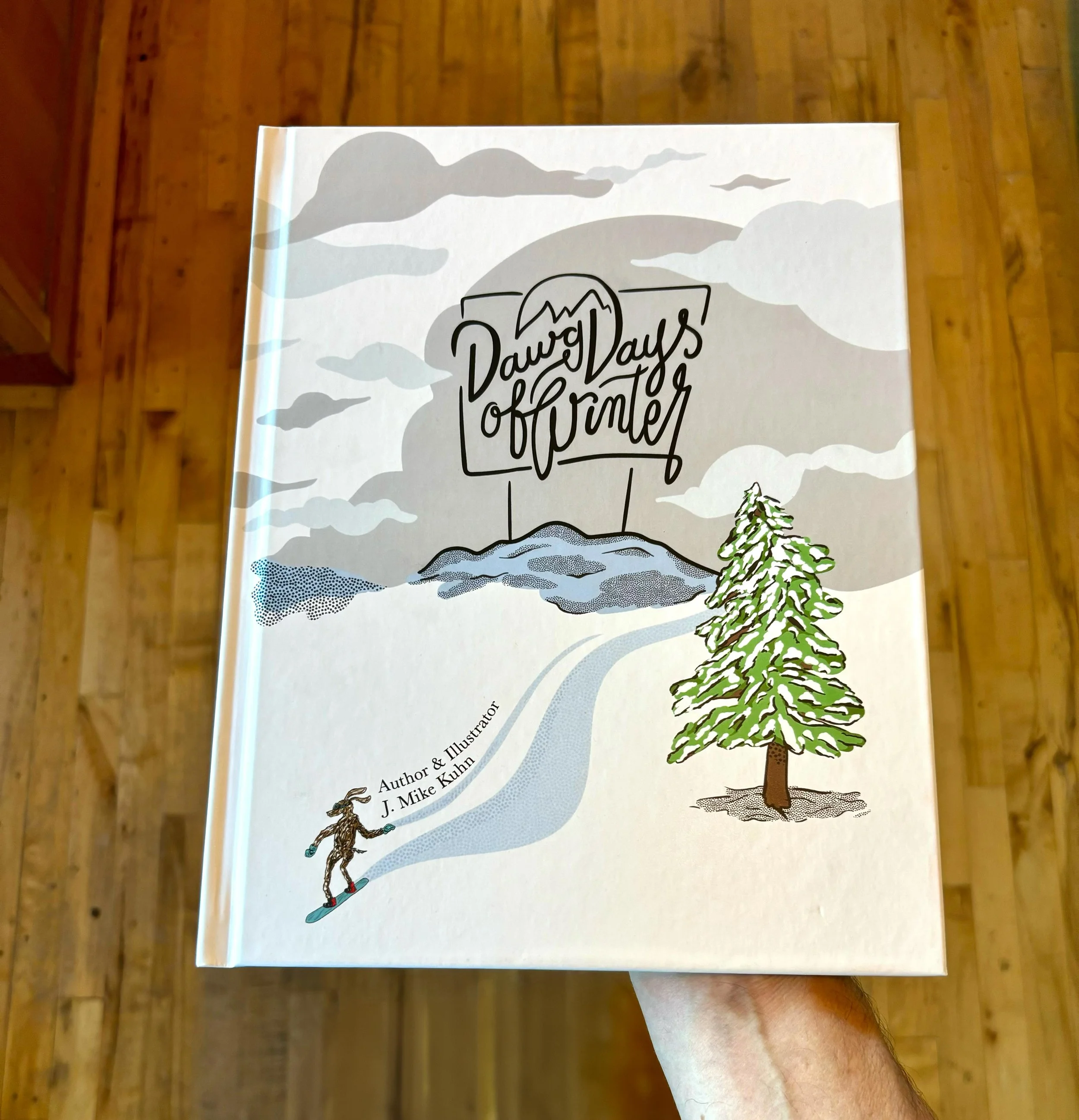 Book titled 'Dawg Days of Summer' with a cover illustration of mountains, clouds, and a pine tree, held in a person's hand over a wooden floor.