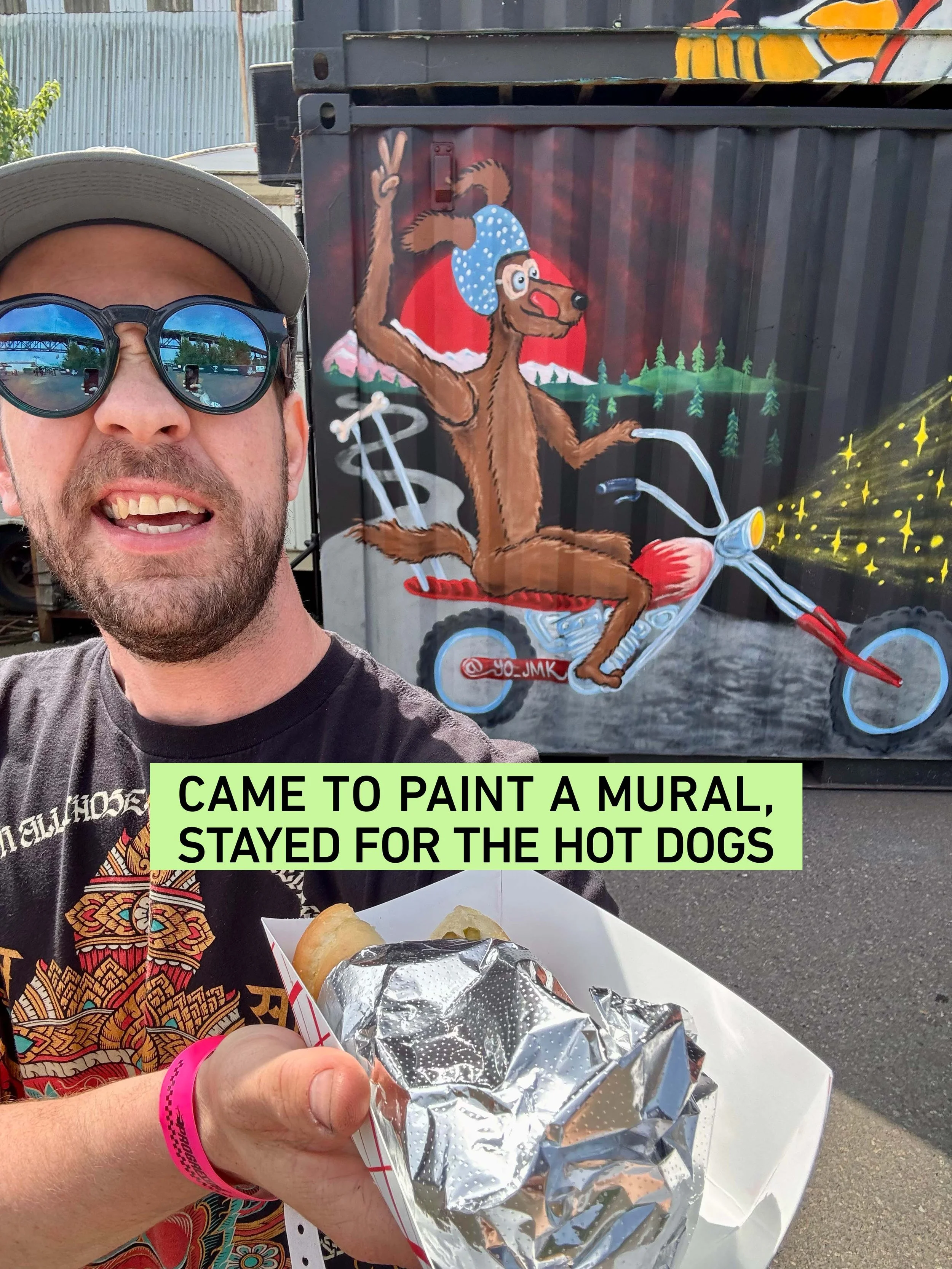 A man wearing sunglasses, a gray cap, and a black T-shirt with colorful designs, holding a hot dog wrapped in aluminum foil. Behind him is a colorful mural of a cartoon dog riding a motorcycle, making a peace sign with its paw, wearing a blue polka dot bandana and glasses, with a mountainous background and pine trees.