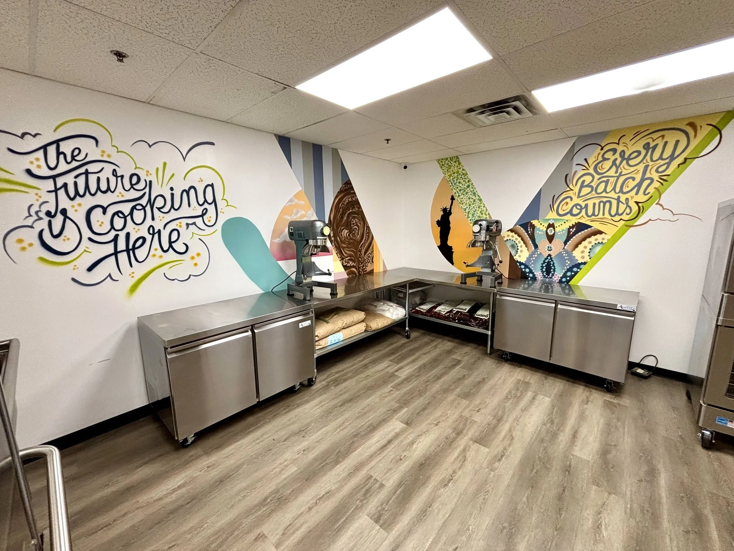 Interior of a commercial or industrial kitchen with a colorful wall mural, stainless steel counters, and a coffee machine.