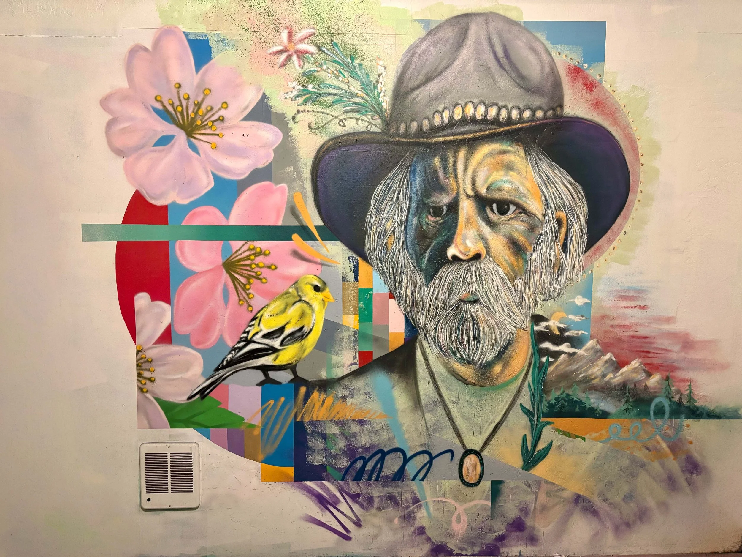 A colorful abstract mural featuring a detailed portrait of an elderly man with a gray beard and a wide-brimmed hat, surrounded by pink flowers, a small yellow bird, and various geometric shapes and patterns.