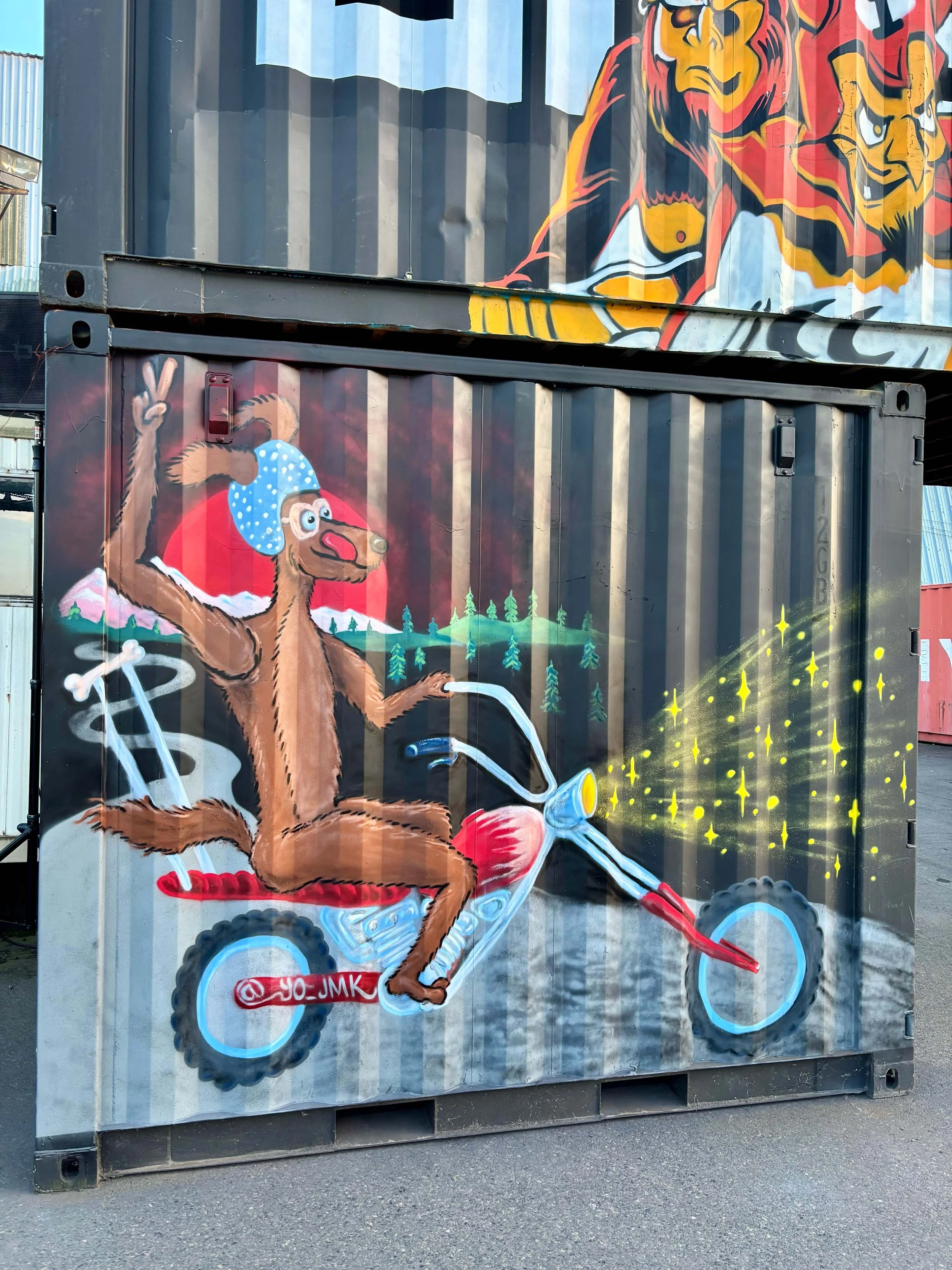 Colorful graffiti mural on a shipping container depicting a dog wearing a blue polka-dotted bandana riding a bicycle with yellow sparkles around. In the background, there is a large mural of a lion and a tiger painted on another container.