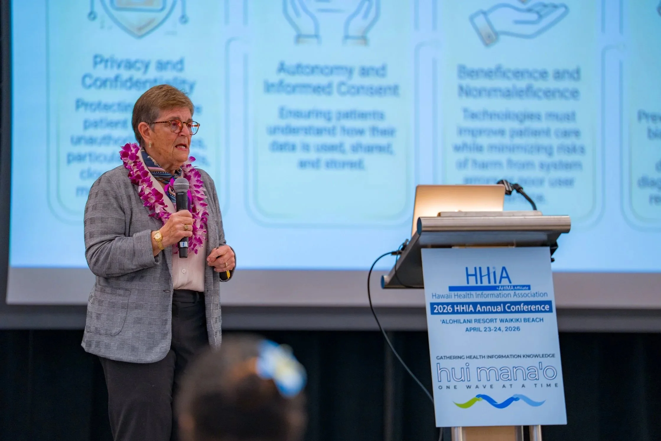A woman with short hair and glasses, wearing a gray checkered blazer and a lei, speaking into a microphone at a conference. Behind her is a large screen displaying conference information and a white podium with the Hawaii Health Information Association logo.