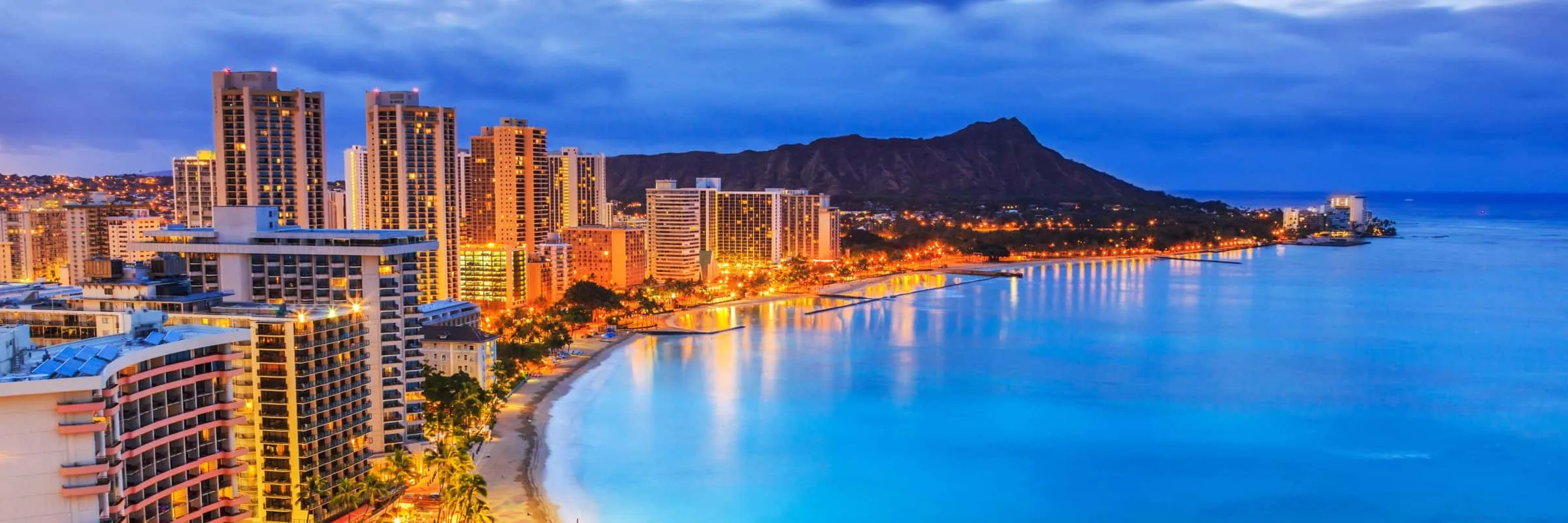 Night view of a city skyline along the coast with tall buildings, streets lit up, and mountains in the background.