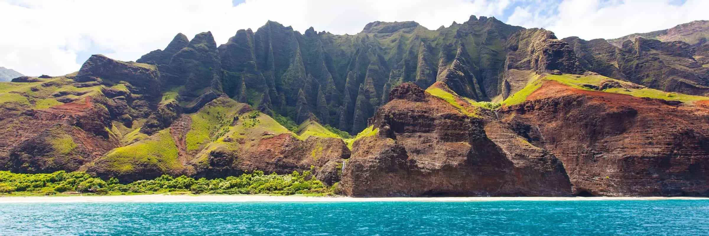 Tropical mountain landscape with rugged green mountains and a shoreline with blue waters in the foreground.