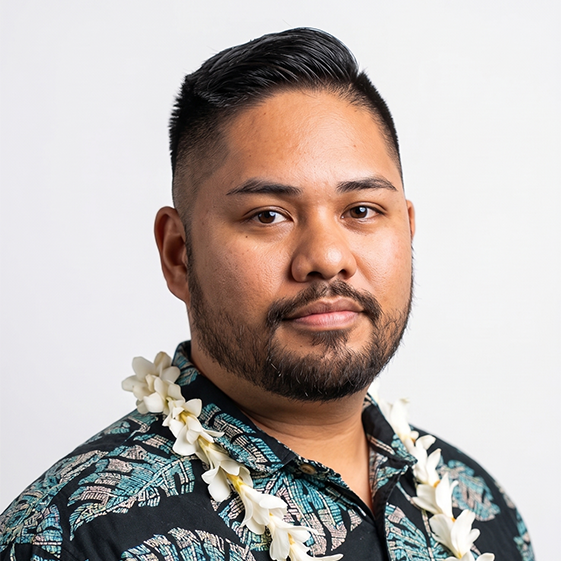 A man with dark hair and a beard wearing a floral shirt and a white lei around his neck, standing against a white background.