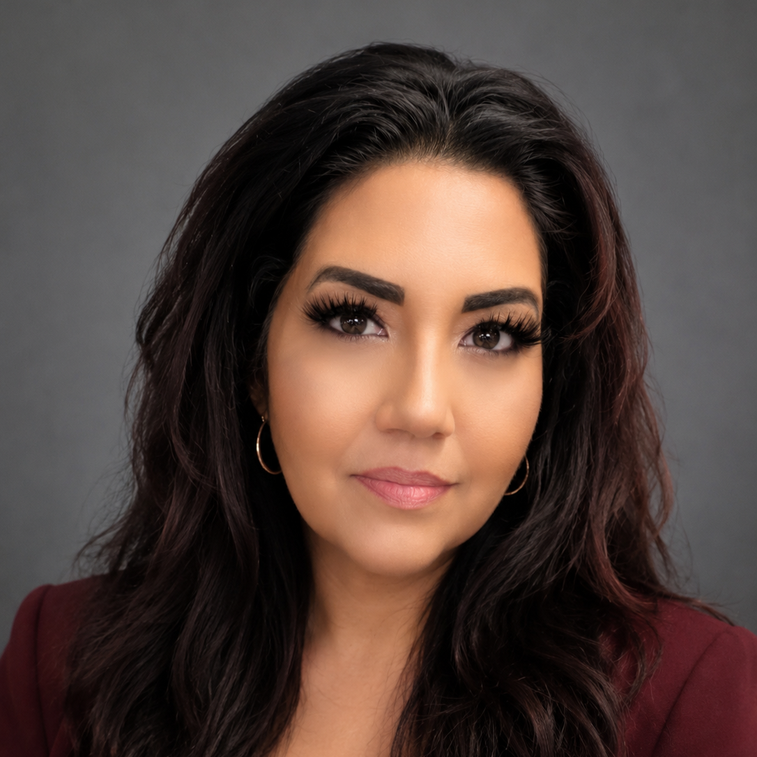 A woman with long dark hair, makeup, and hoop earrings, looking directly at the camera against a gray background.