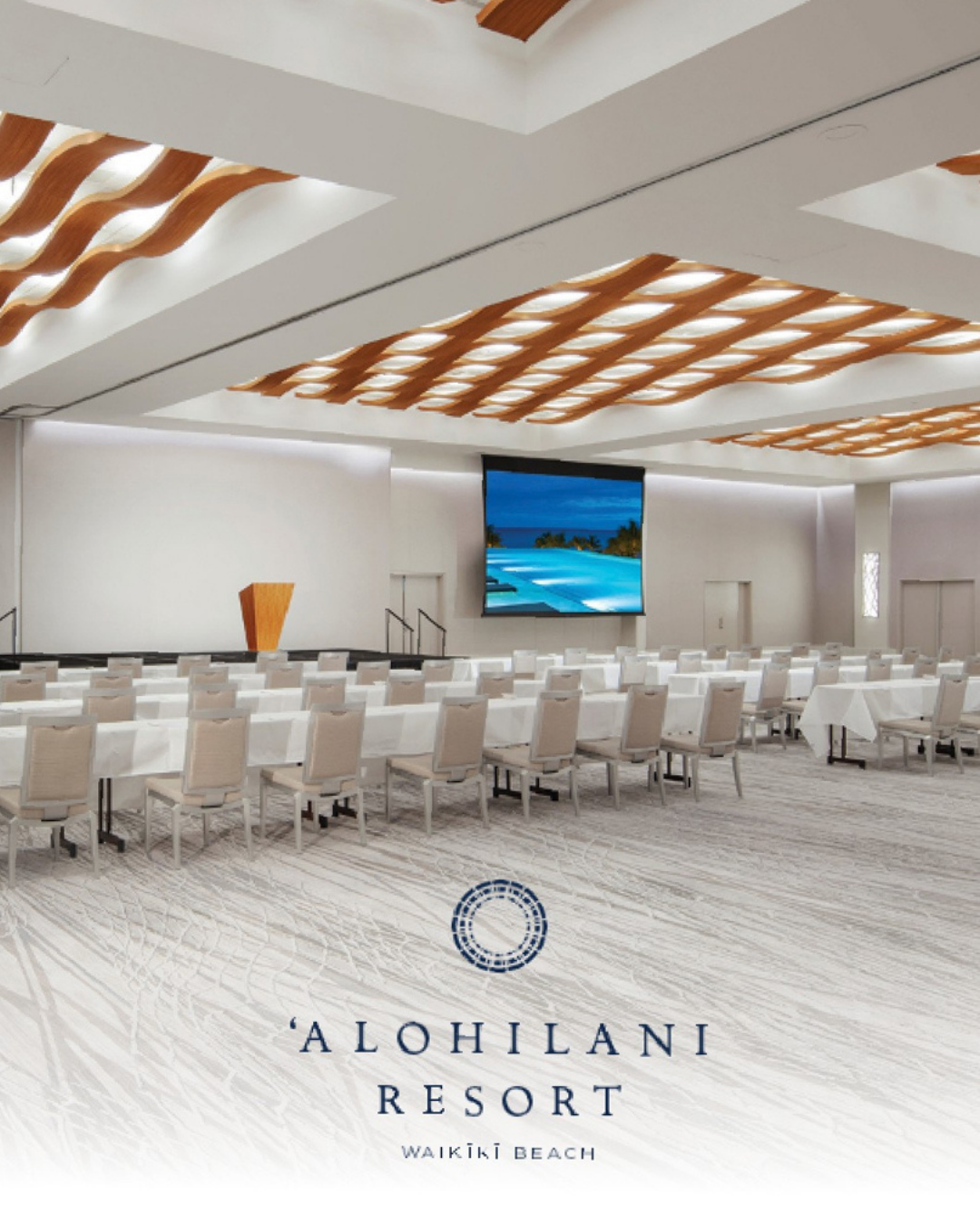 A spacious conference room with rows of white chairs and tables, a large screen displaying a beach scene, and decorative wooden wave patterns on the ceiling at Alohilani Resort, Waikiki Beach.