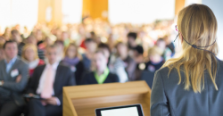 A woman speaking at a podium in front of an audience in a large, well-lit room or hall