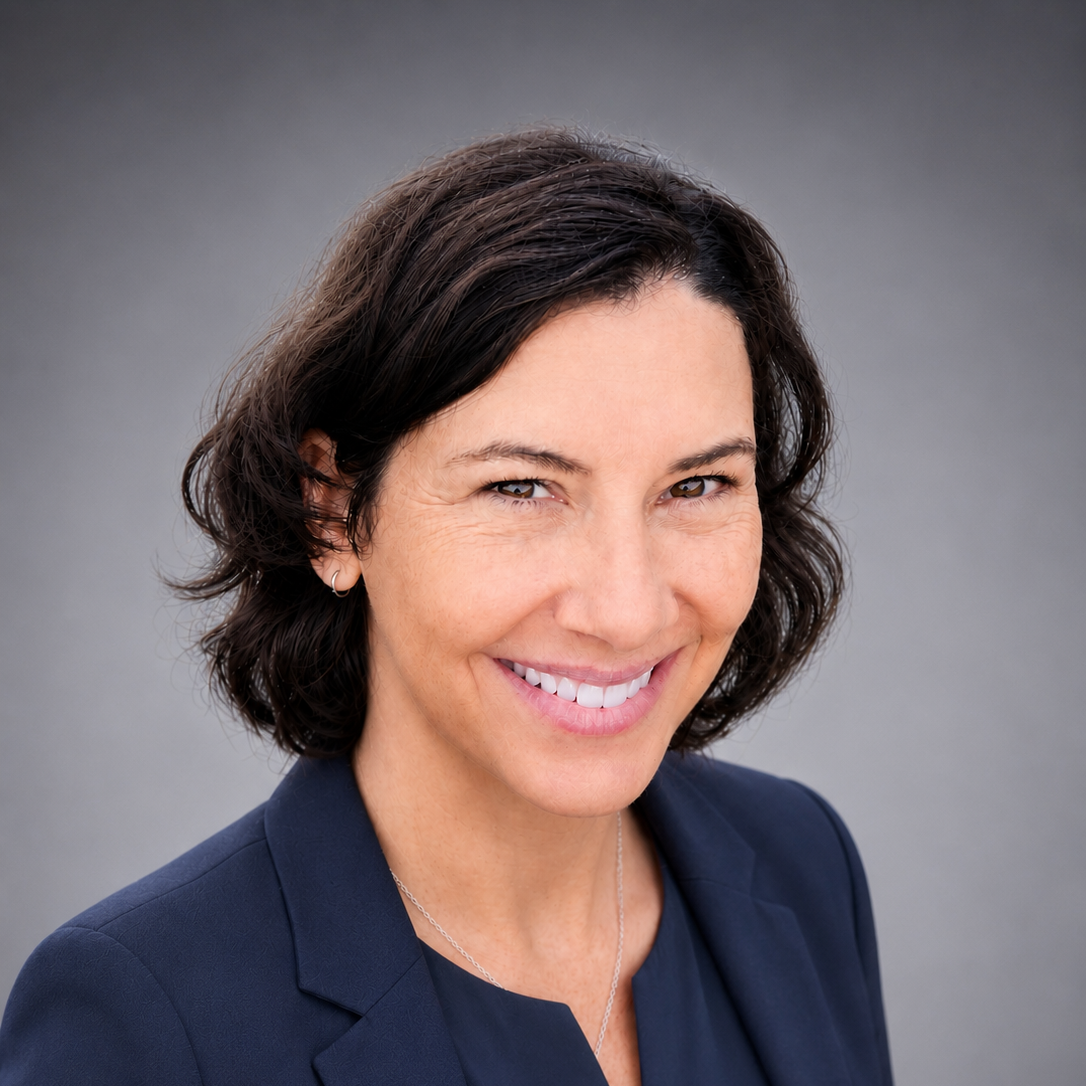 Smiling woman with dark wavy hair wearing a navy blazer and a silver necklace against a gray background.