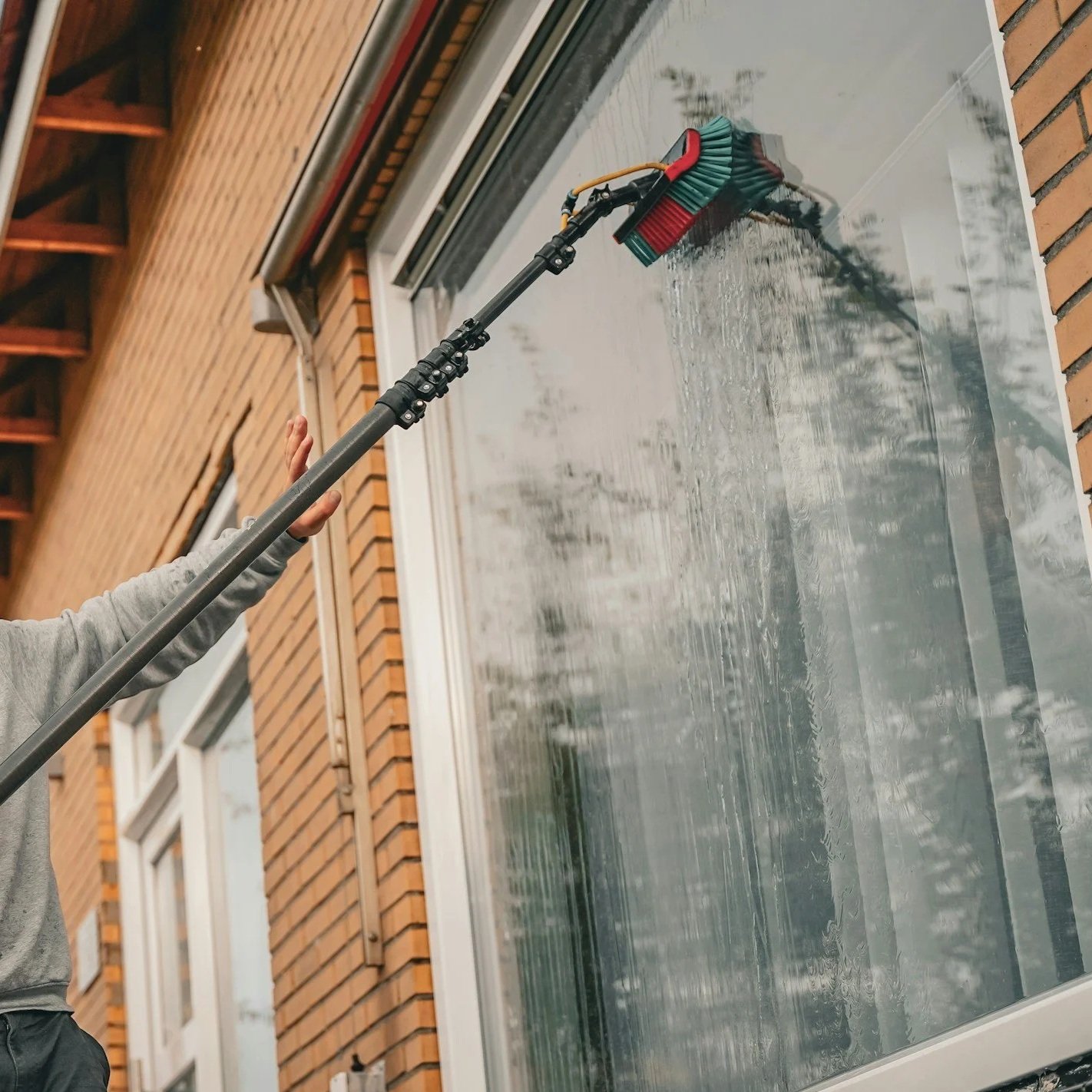A person using a long-handled cleaning tool to clean a large glass window on a brick building