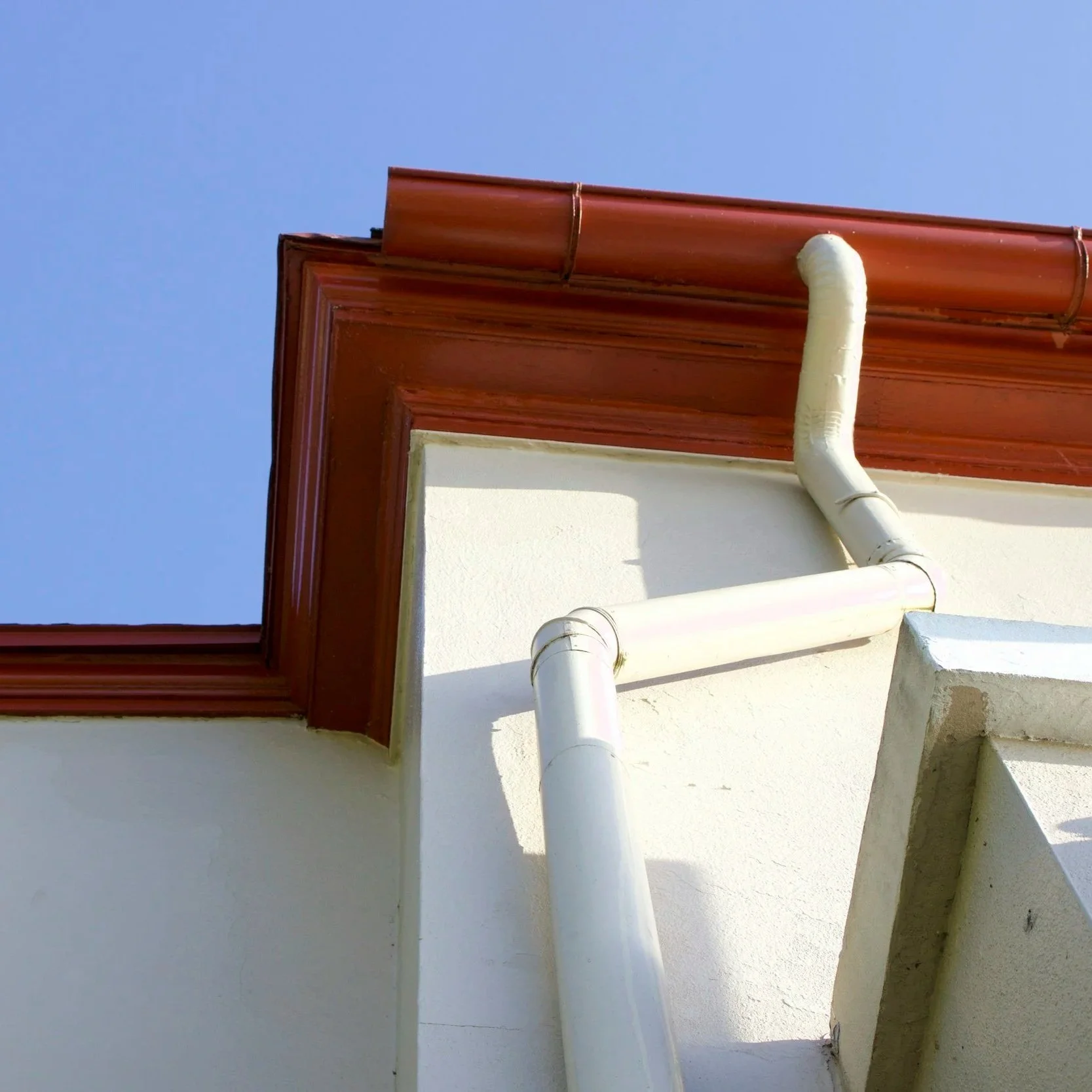Close-up of a house corner with a brown roof overhang, white vertical drain pipe, and part of a staircase with concrete stairs against the white wall, under a clear blue sky.