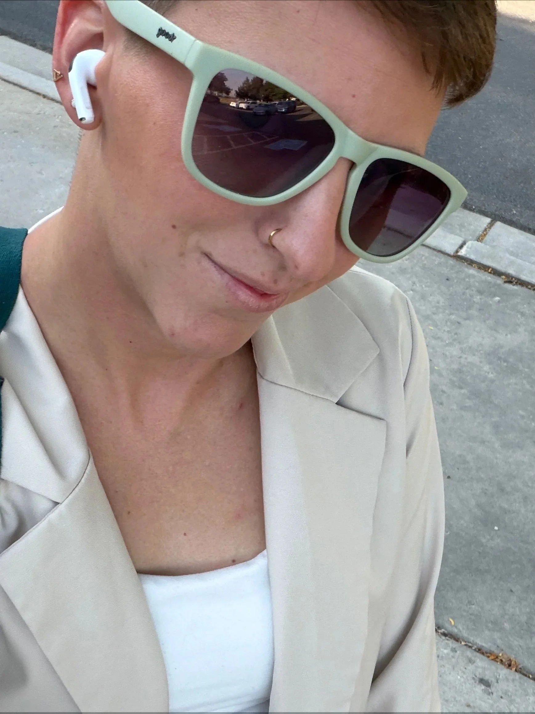 Close-up of a woman with short brown hair wearing light green glasses, white makeup, a gold nose ring, white earbuds, a white top, and a beige blazer standing on a sidewalk.