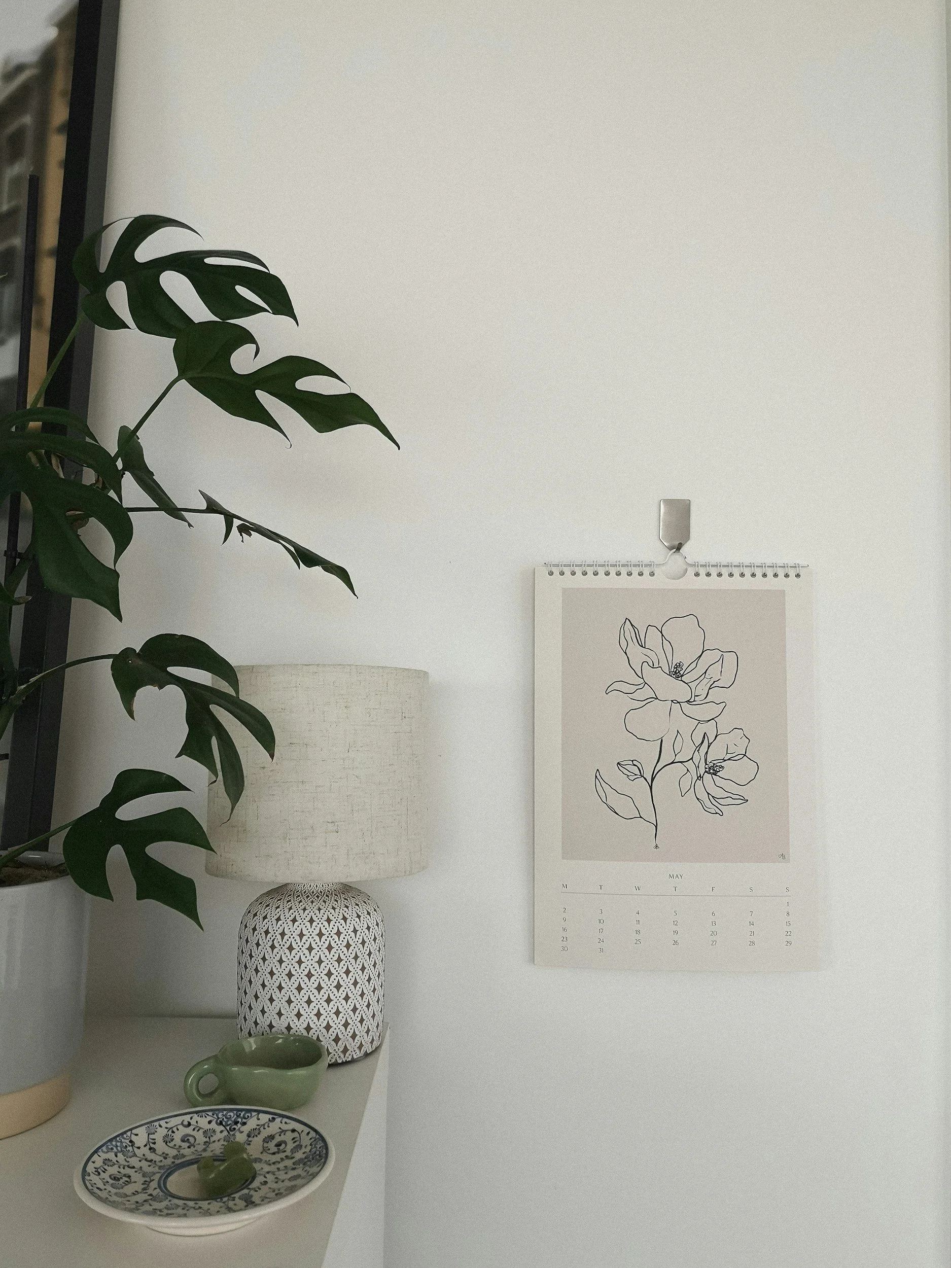 Decorative vignette with indoor plants, a ceramic table lamp, a green mug, and a black and white floral calendar on a white wall.