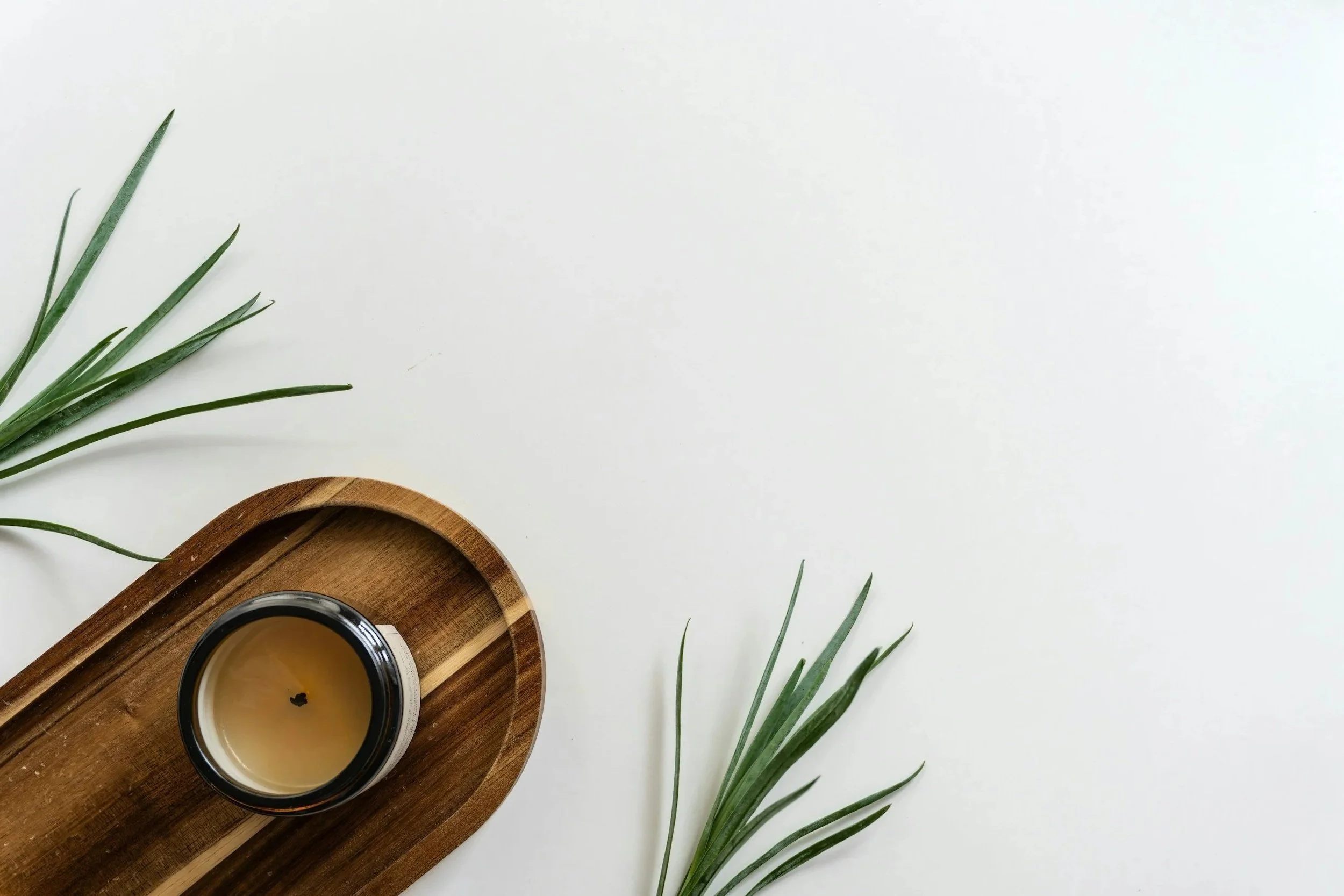 Top-down view of a wooden tray holding a black-capped glass of tea, on a white surface, with green foliage on the left and bottom edges.