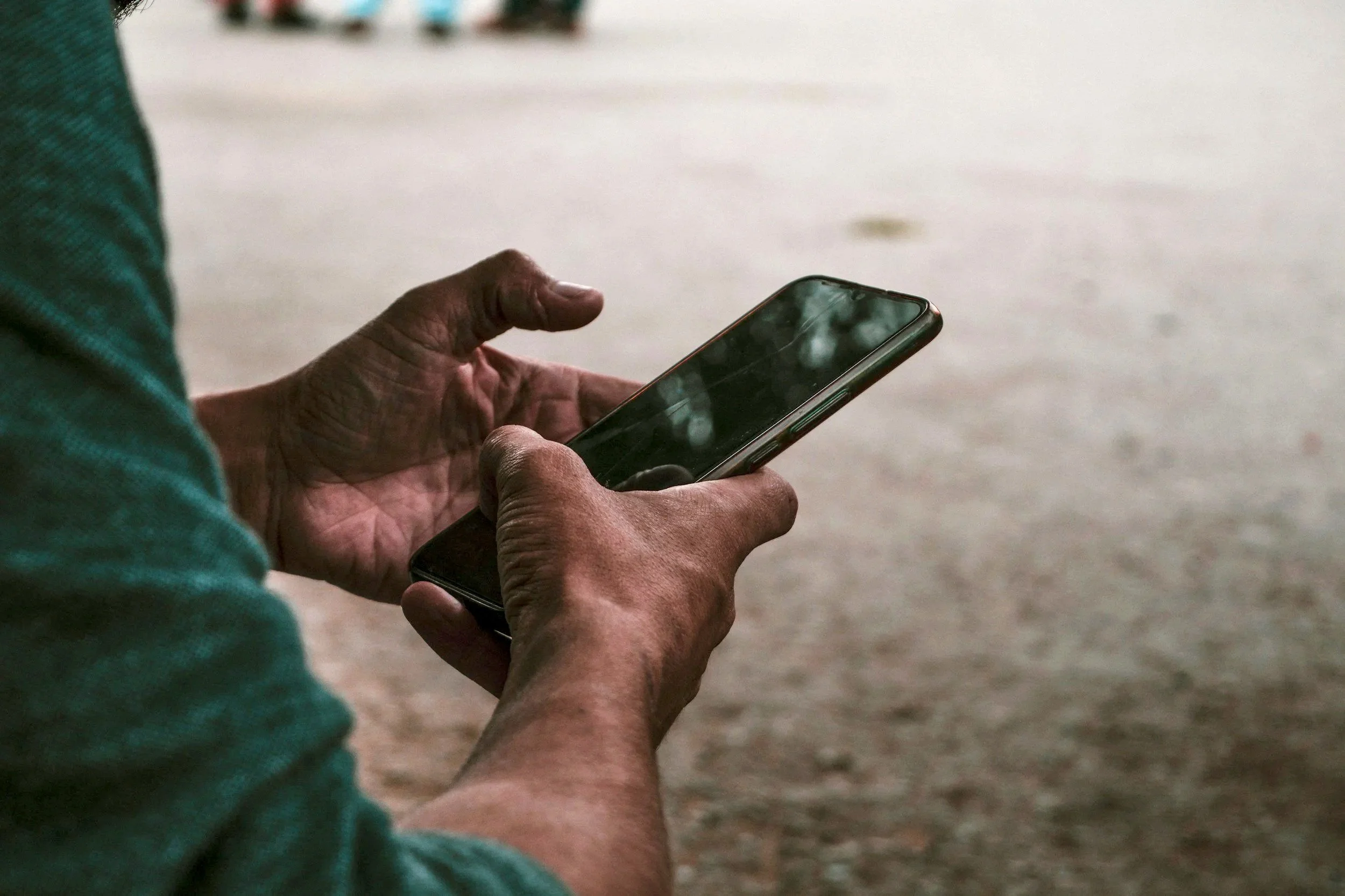 Close-up of a person with dark skin using a smartphone outdoors, with a green sleeve partially visible on their arm and a blurry background.