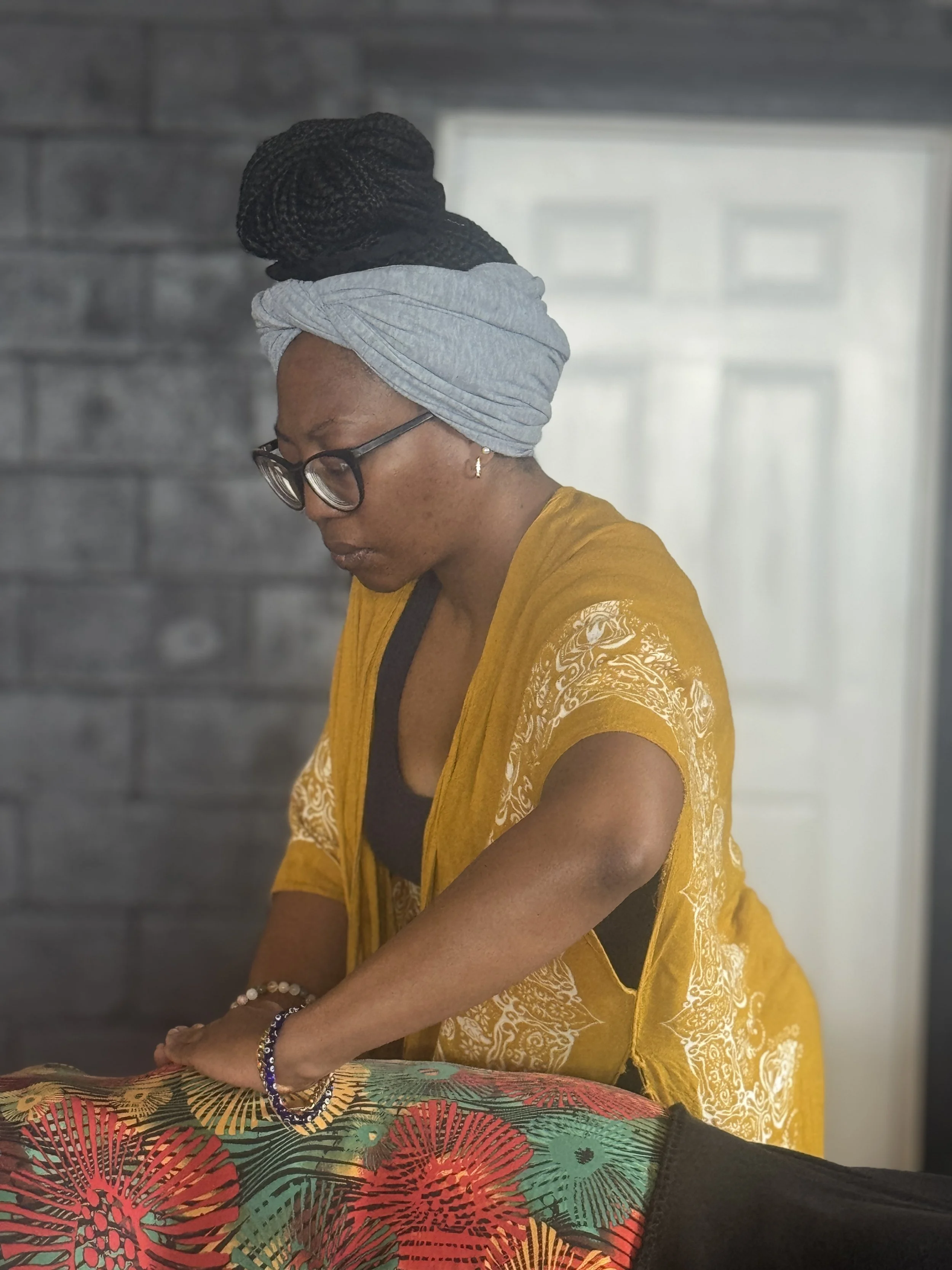 A mahogany brown-skinned woman wearing glasses, a gray headwrap, and a yellow garment with white patterns, her hair tied in braids, and jewelry including a bracelet and earring, standing indoors against a gray wall and a window in the background.