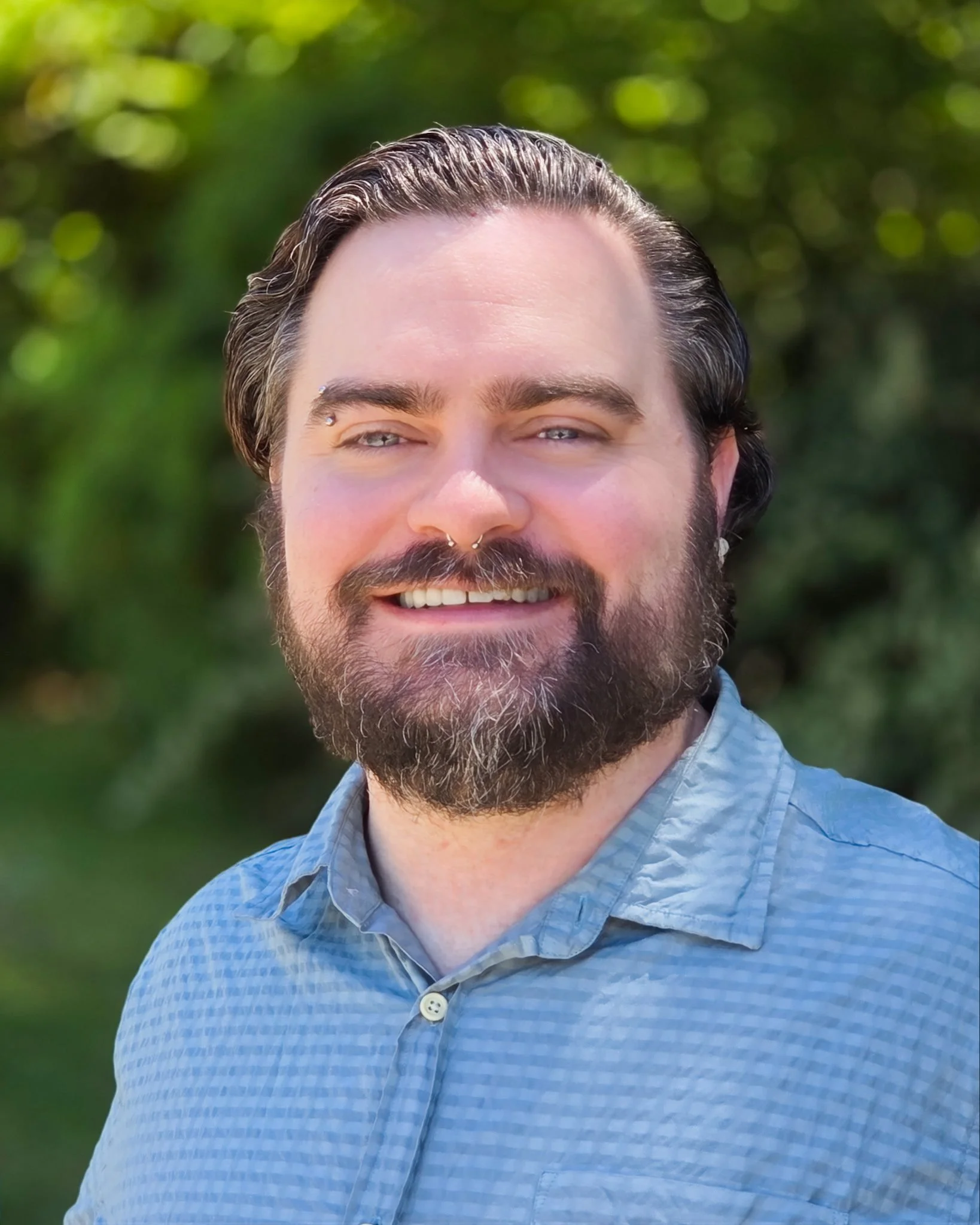 Close-up of a smiling man with a beard, mustache, and eyebrows, wearing a light blue checkered shirt with a collar, outdoors with green trees and blurred background.