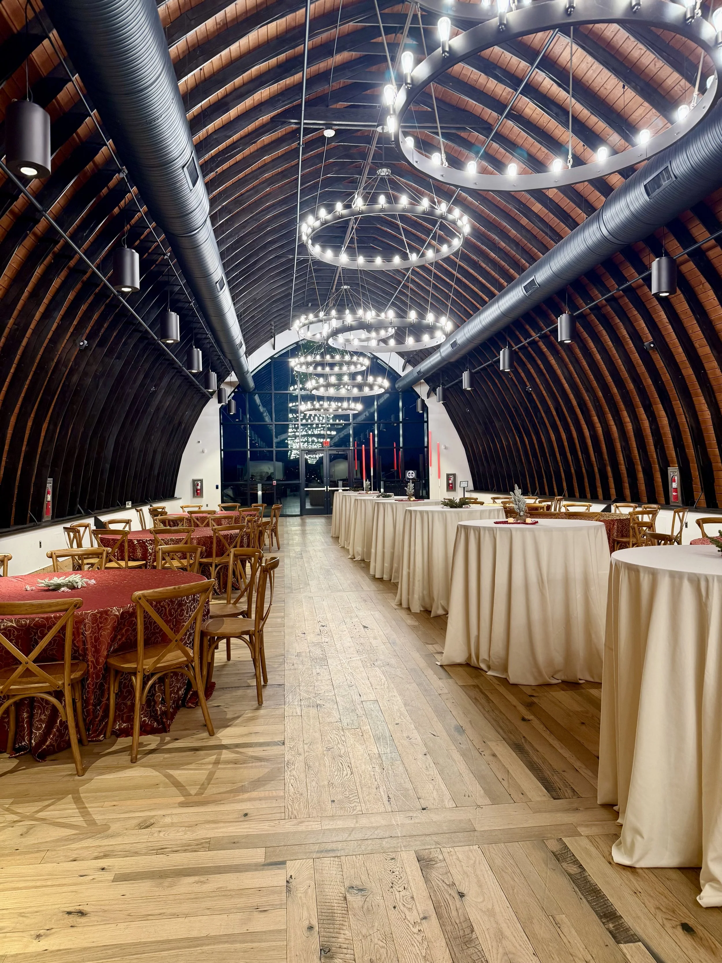 Interior of a large, elegant hall with arched wooden ceiling, black hanging cylindrical lights, and circular chandelier lighting fixtures. There are tables with cream-colored tablecloths and floral centerpieces, surrounded by wooden chairs. The floor is made of light-colored wood planks, and there are glass doors at the far end.