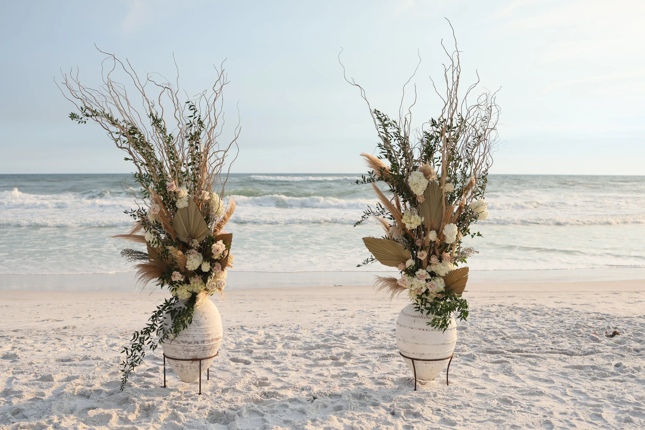 Two floral arrangements on white vases with driftwood and greenery, set on a sandy beach with ocean waves in the background.