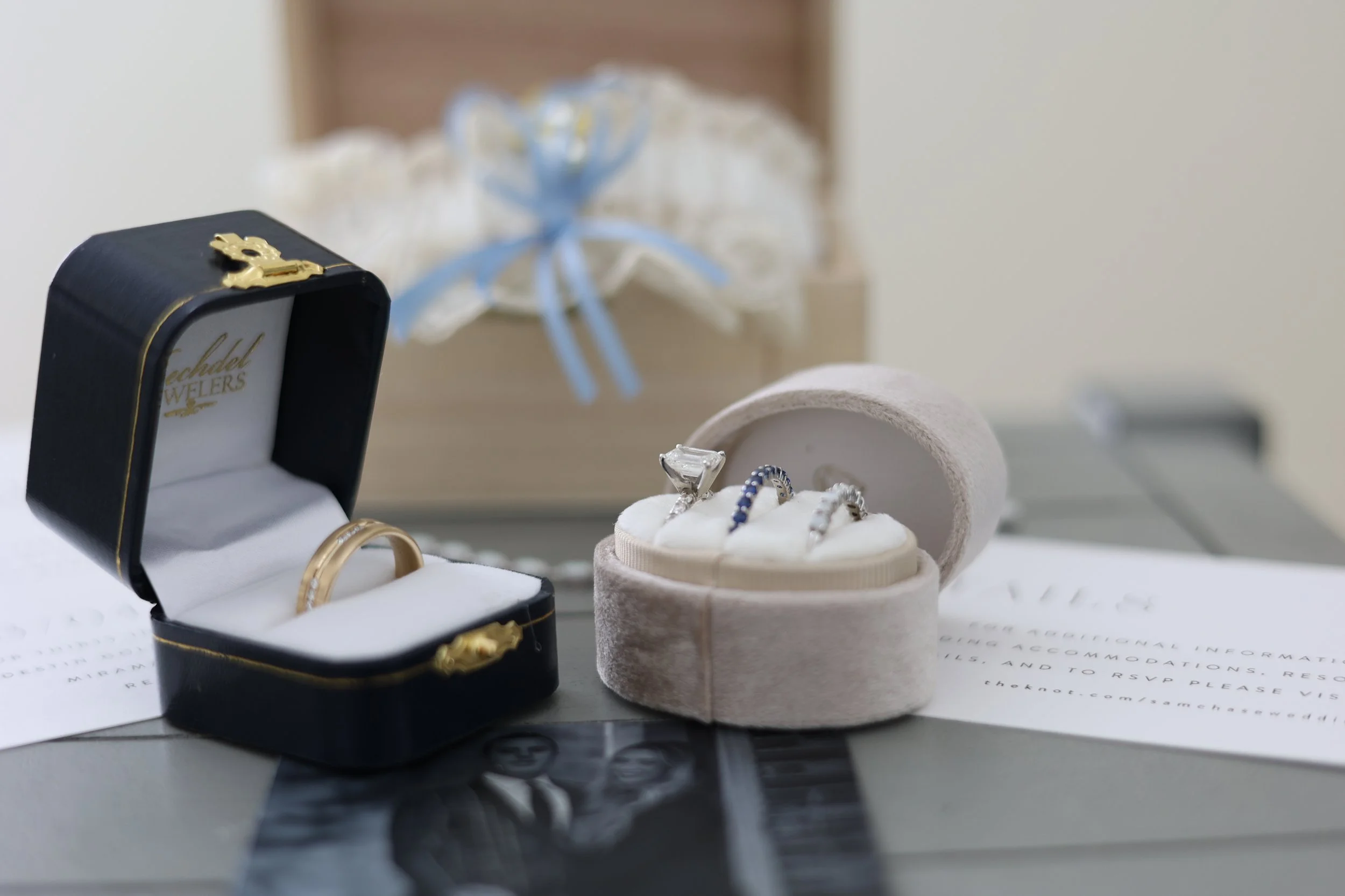 A black jewelry box with gold embellishments and a logo that says 'Rebecca Jewelry' contains gold rings. Behind the jewelry box, there is a small beige ring box with a textured surface holding a diamond ring and several other rings with gemstones. In the background, there is a gift wrapped with clear plastic and a blue ribbon, along with white papers and a document with text.