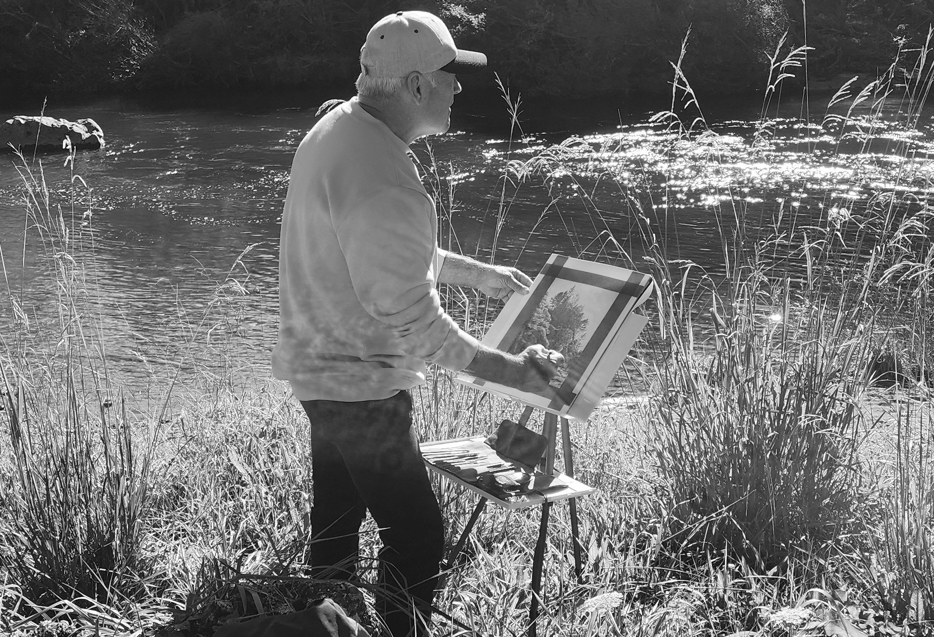 An elderly man wearing a cap and a light-colored shirt stands outdoors, painting on a canvas set on an easel, beside a river with tall grasses and rocks, capturing the natural landscape.
