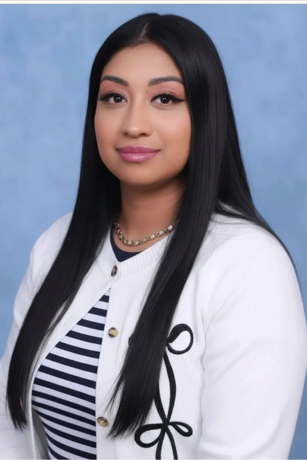 Professional portrait of a young woman with long black hair, wearing a white cardigan with black embroidery, a striped shirt, a pearl necklace, and a gentle smile, against a light blue background.