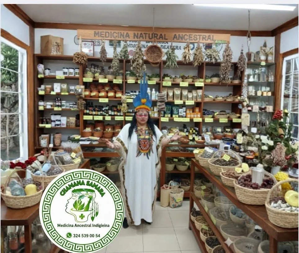 Mujer vestida con traje indígena en una tienda de productos naturales y medicinales ancestrales, con estanterías llenas de hierbas, especias y otros ingredientes, y un cartel que dice 'Medicina Natural Ancestral'.