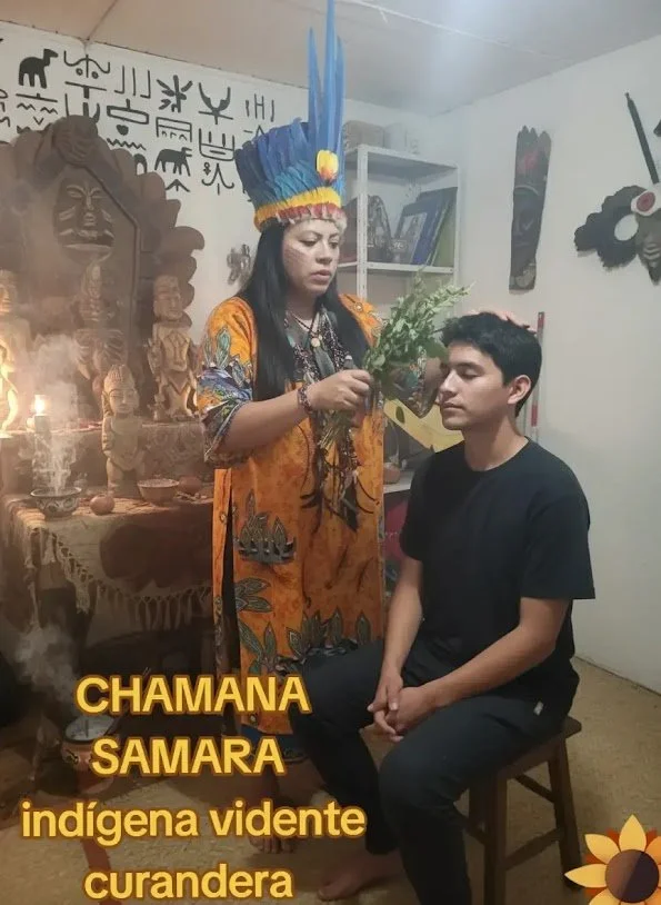 Una mujer indígena vestida con un atuendo tradicional y un tocado de plumas azules, realiza un ritual chamánico con un hombre sentado en una silla. En el entorno hay símbolos y objetos culturales, y letras en jeroglíficos en la pared, además de imágenes de arte indígena y una figura tallada en madera.
