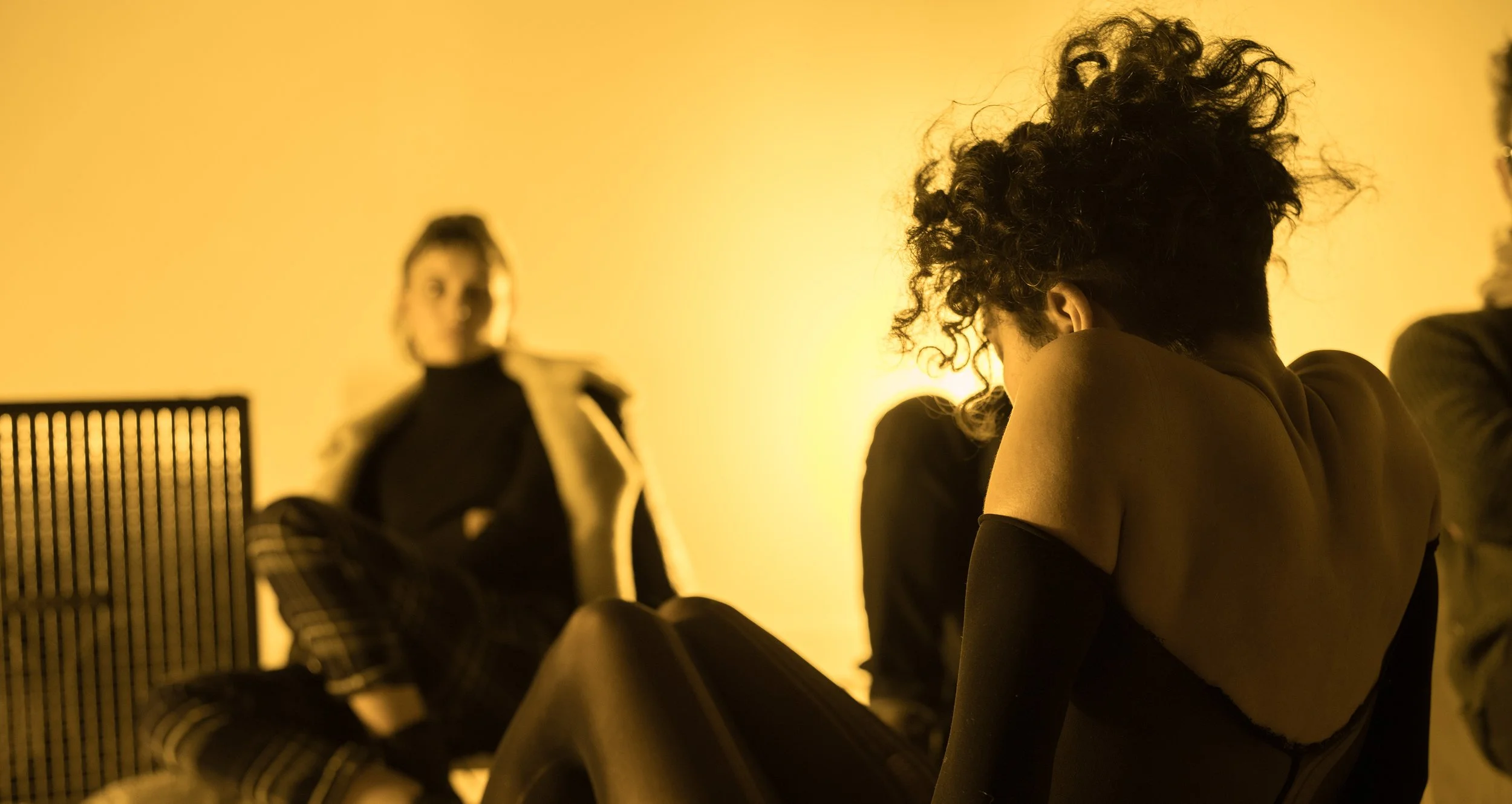A performer seen from the side with her back tense, a yellow setting, and a woman watching her, seated in front of her