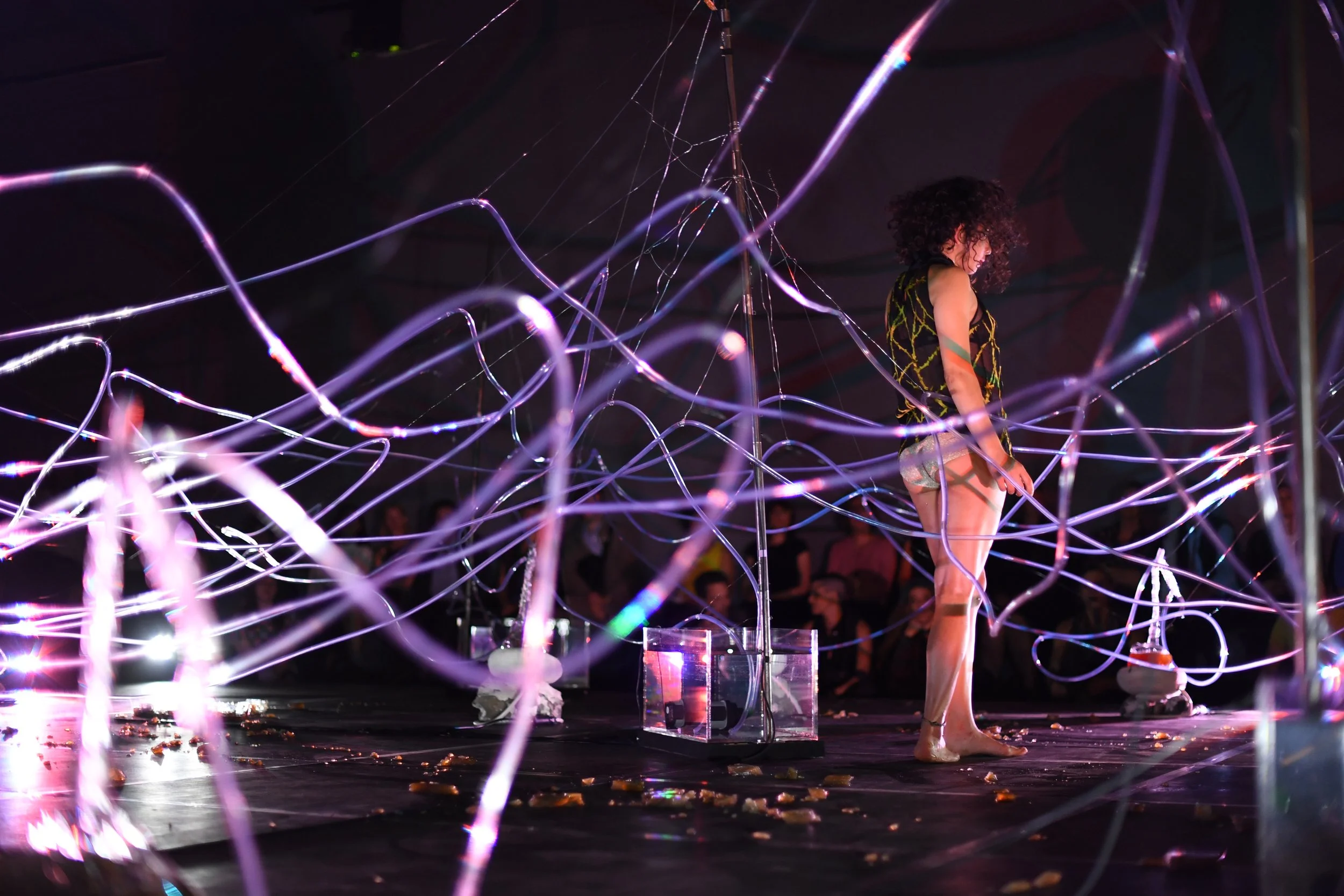 standing dancer performing Basic Transmutation- Alien/Migration, in an installation of transparent tubes and distillers