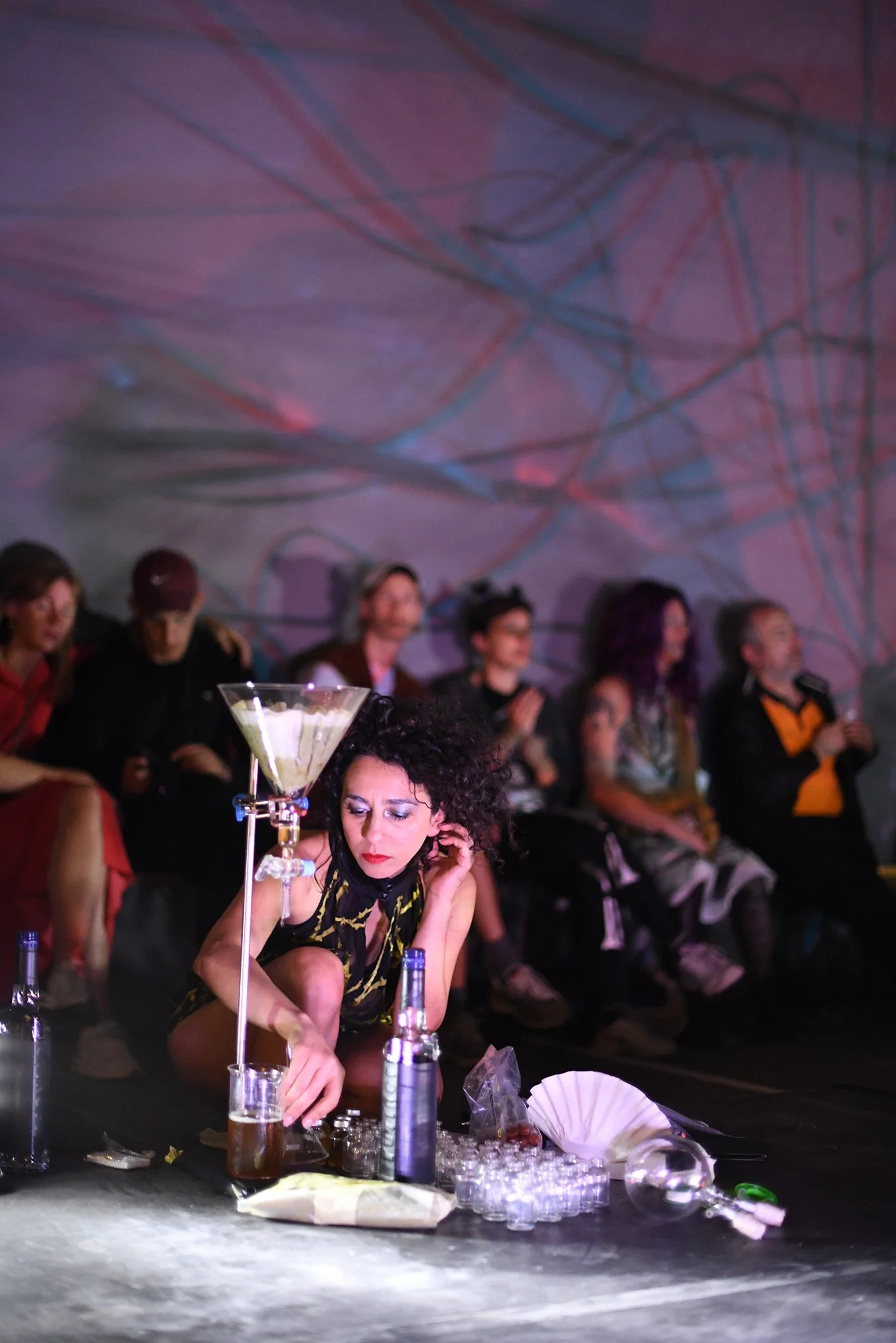 A dancer crouching in front of a percolator, with the audience behind colorful remnants from the tubes of the installation Basic transmutation -Alien/Migration*,