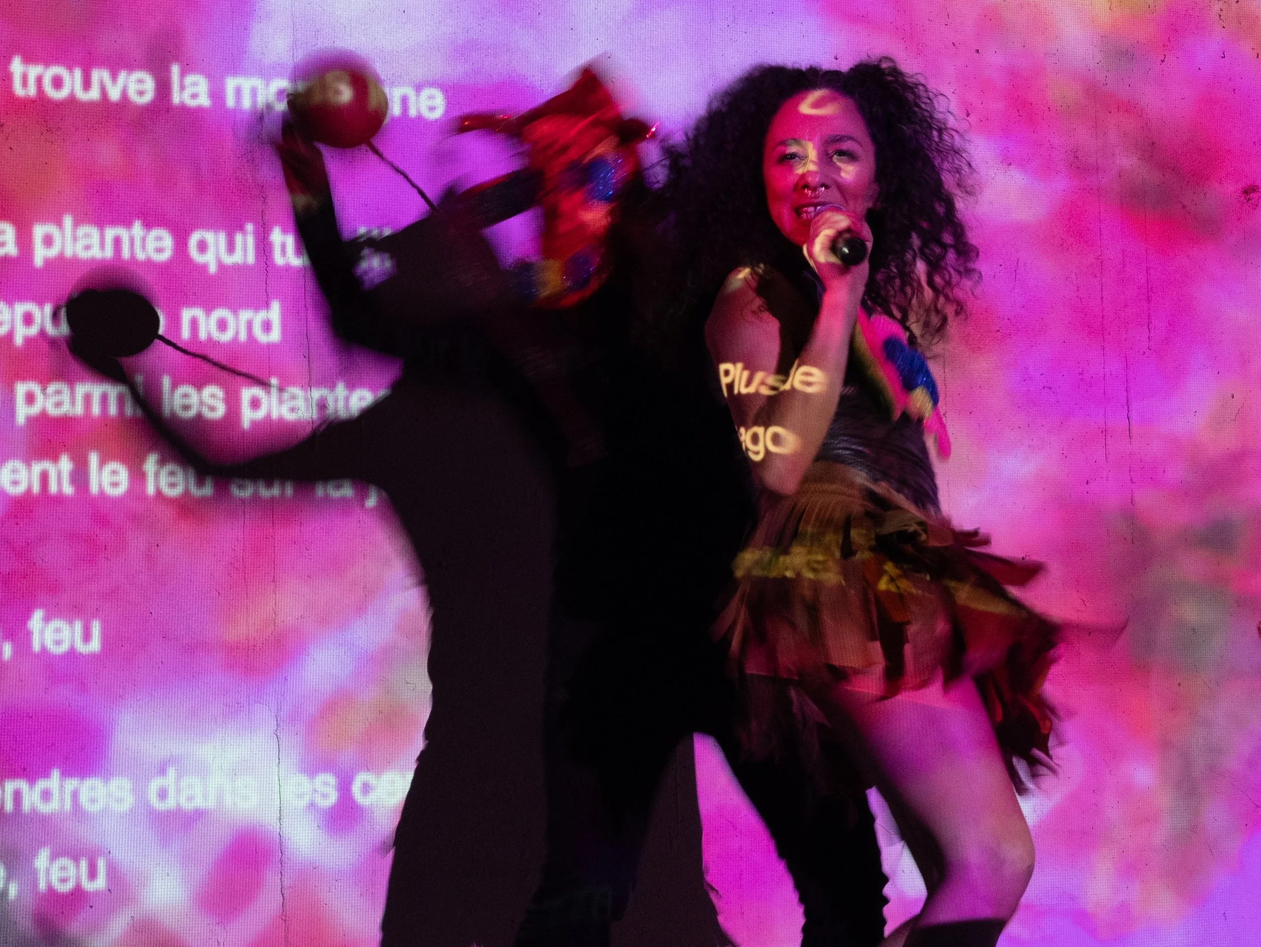 a dancer with a microphone in her hand in front of a projection of pink and red colors, alongside another performer wearing a carnival bull mask and holding maracas.