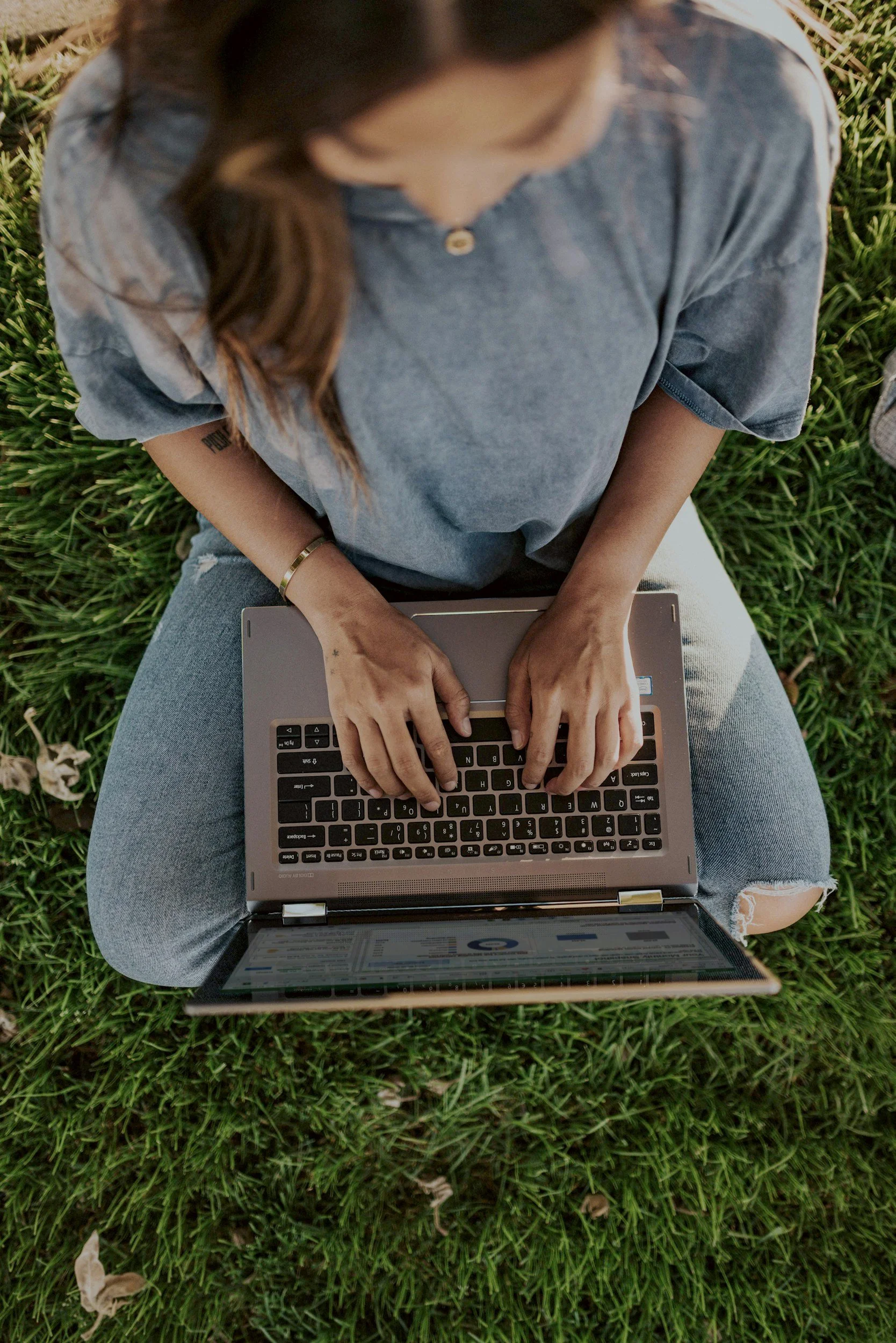 Person sitting on grass with a laptop on their lap, typing on the keyboard, wearing a gray shirt and ripped jeans.