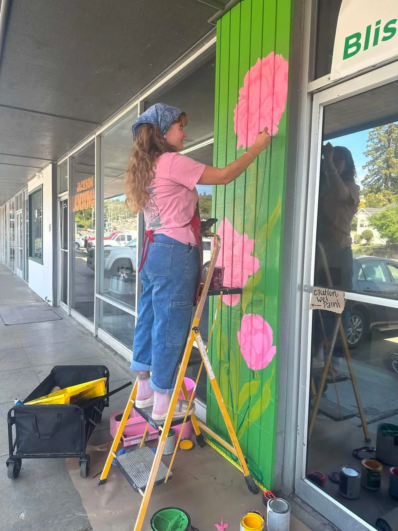 Clare Toraya joyfully working on an exterior mural of botanicals. This mural was covering an old, corrugated wood surface, a complicated surface to work on. She paints vibrant, eye catching flowers with weaving stems for a Thrift Store in Jackson CA