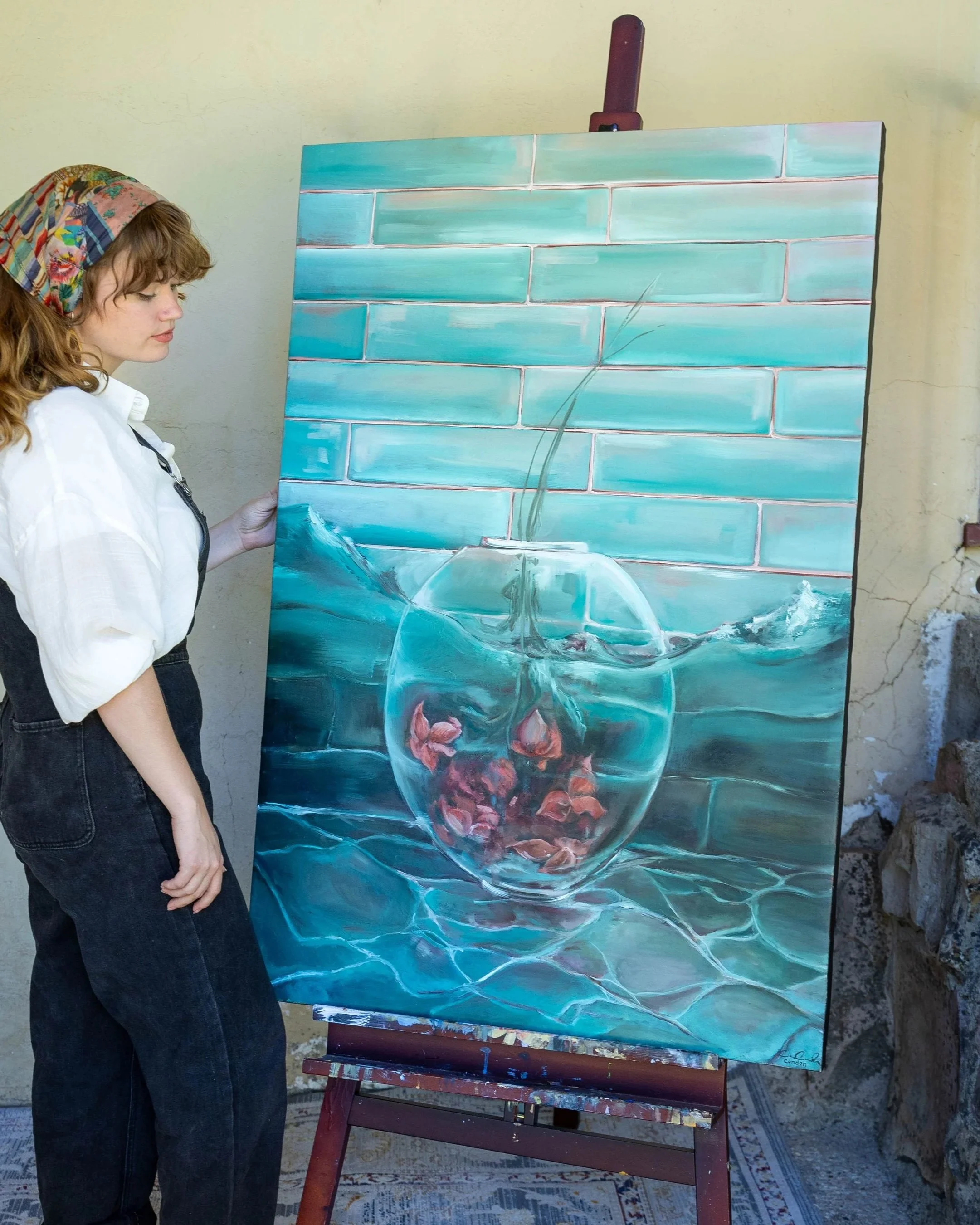 A young woman with curly, reddish-brown hair wearing a colorful headscarf, white shirt, and black pants, stands next to an easel with a painting of a teacup filled with pink roses and a green-and-white tiled wall in the background.