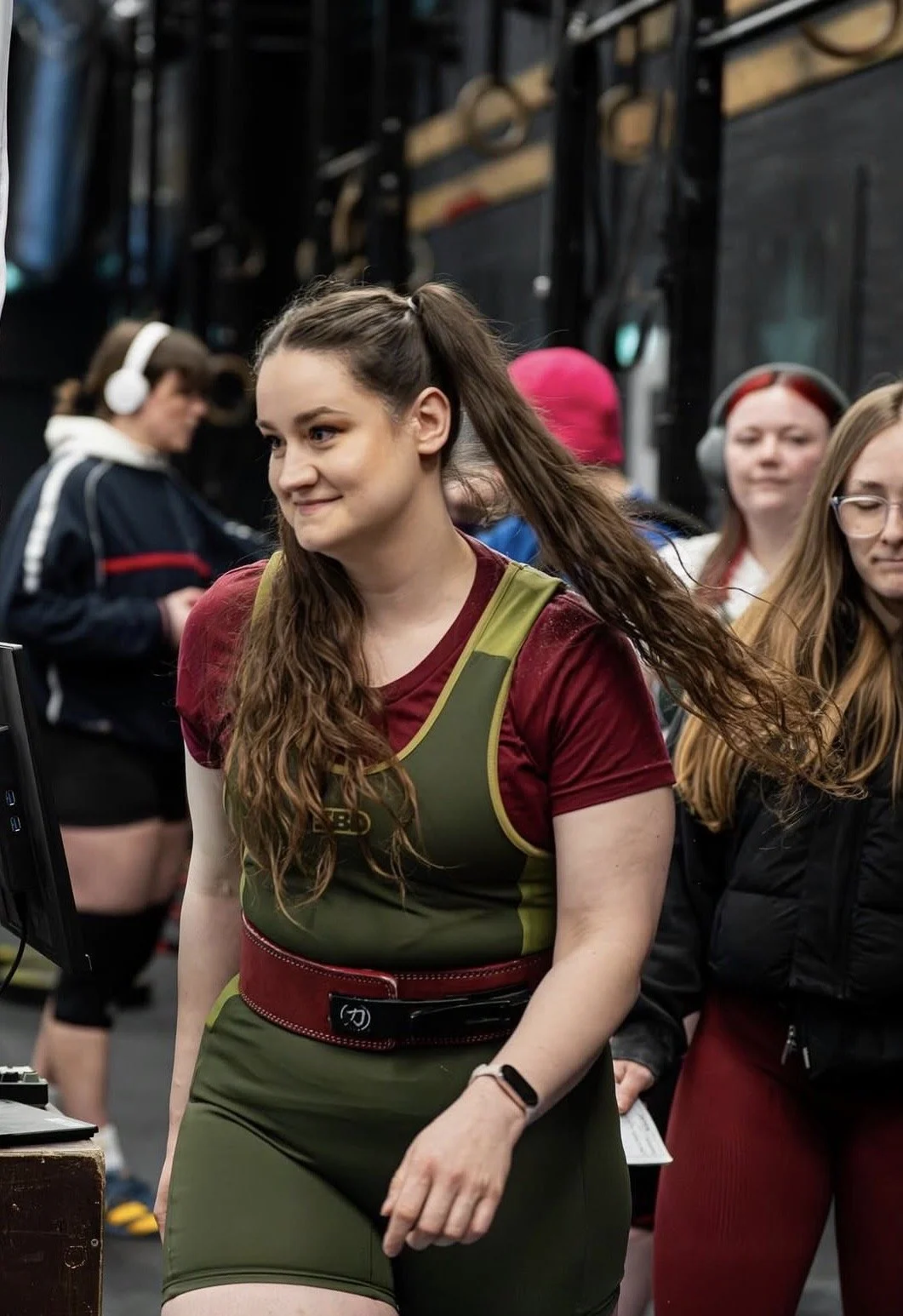 A woman with long brown hair in a ponytail wearing a green and red workout outfit at a gym, surrounded by other women, some wearing headphones.