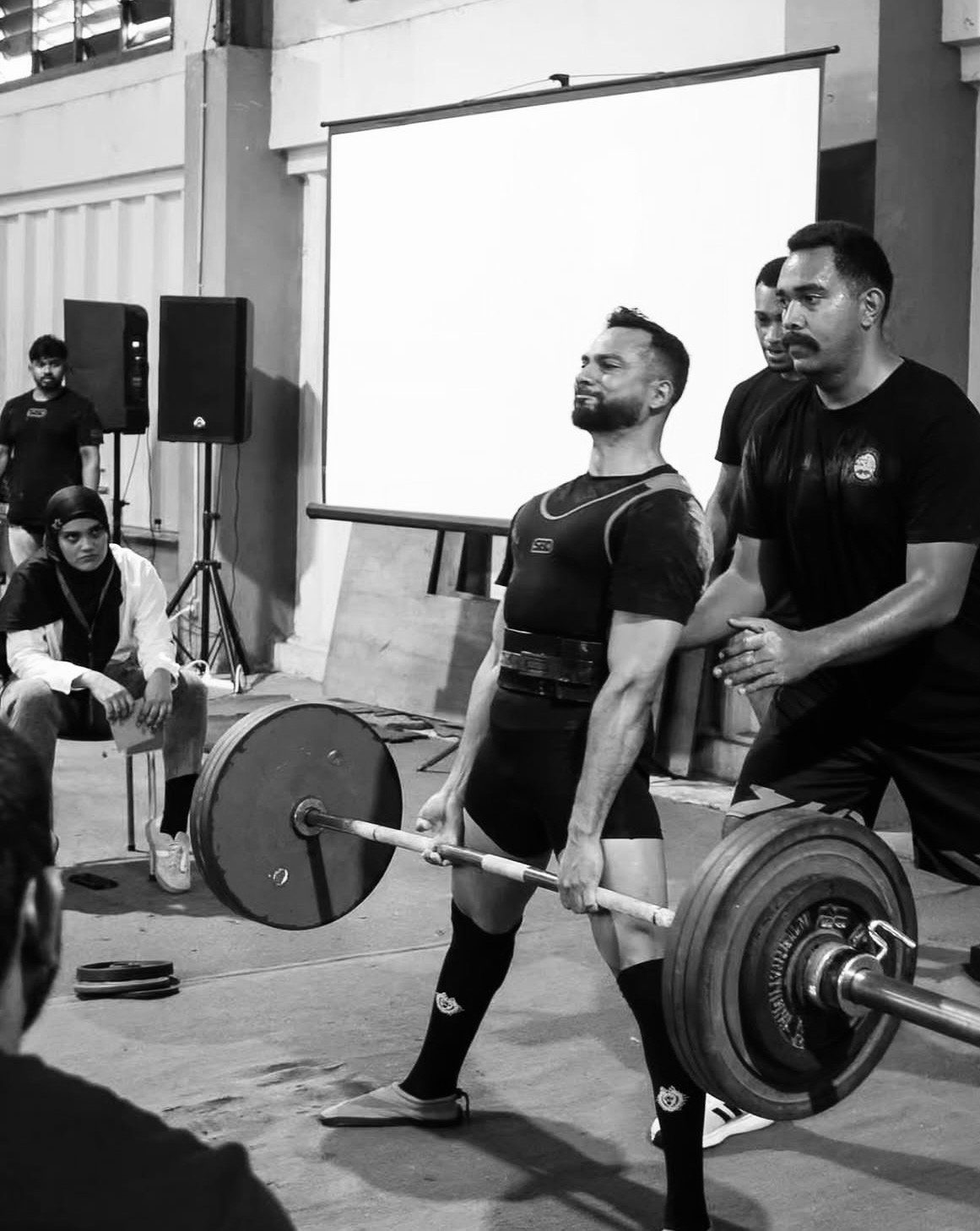 A man in a weightlifting uniform is lifting a barbell with weights. Two other men are assisting or spotting him, one standing behind him and another standing nearby. Several people are watching, including a woman sitting on a chair and a person seated on the floor in the foreground. A large blank screen and speakers are visible in the background.