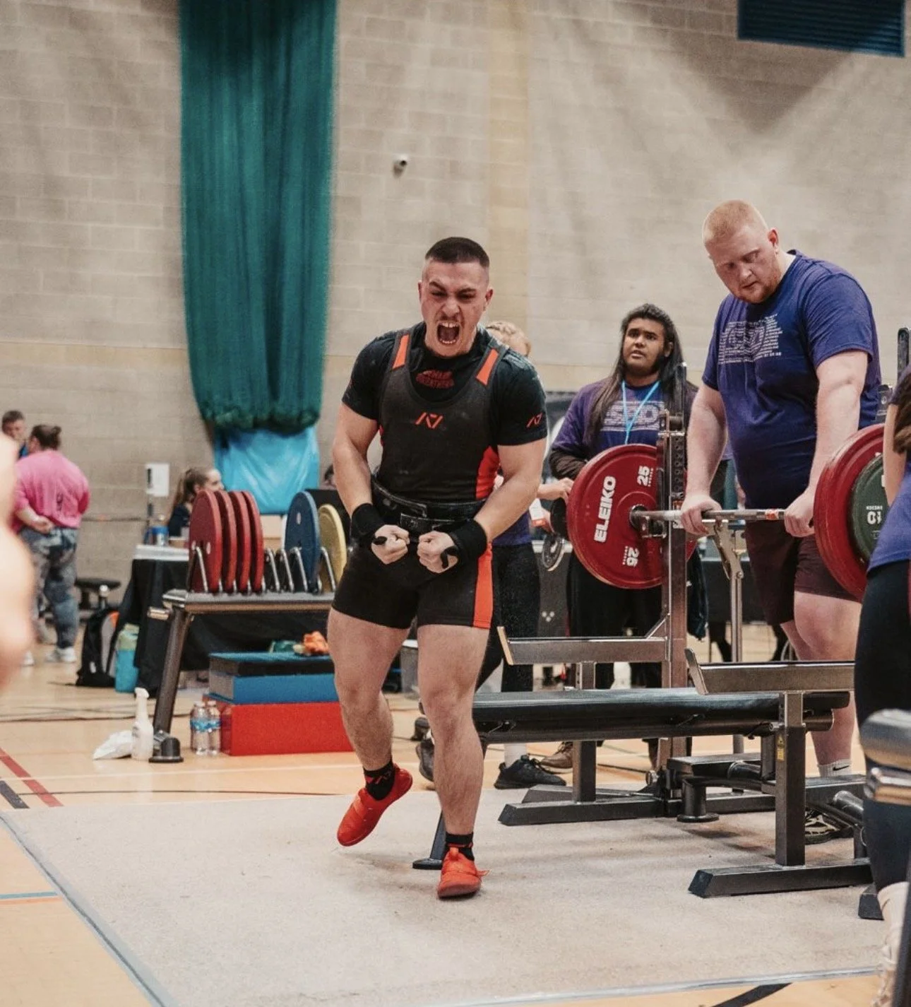 A powerlifter in a black and red outfit celebrating after a lift at a competition, with other people and equipment nearby in an indoor gym or competition setting.
