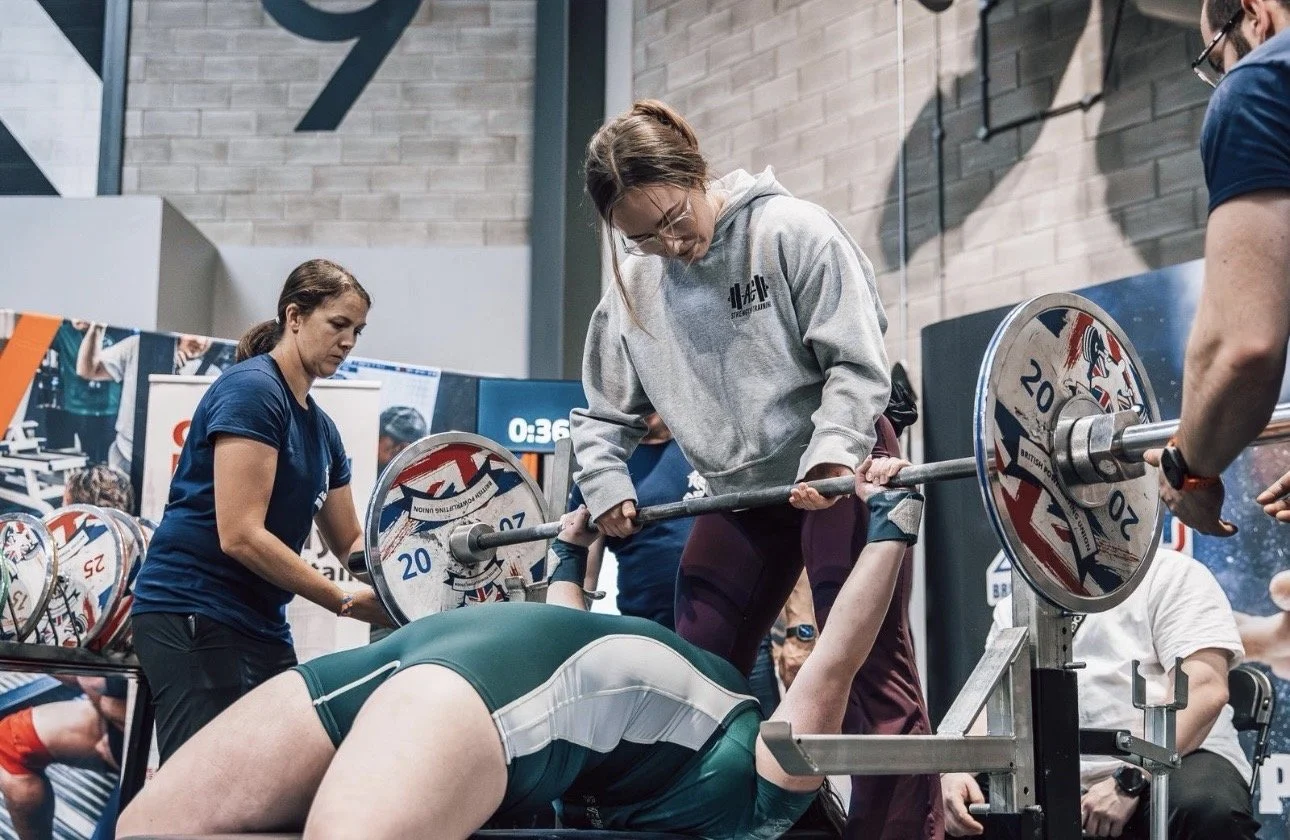 A woman in green and white workout gear is lifting a barbell with weights while another woman in a gray hoodie assists, and a spotter in blue is nearby at a powerlifting competition.