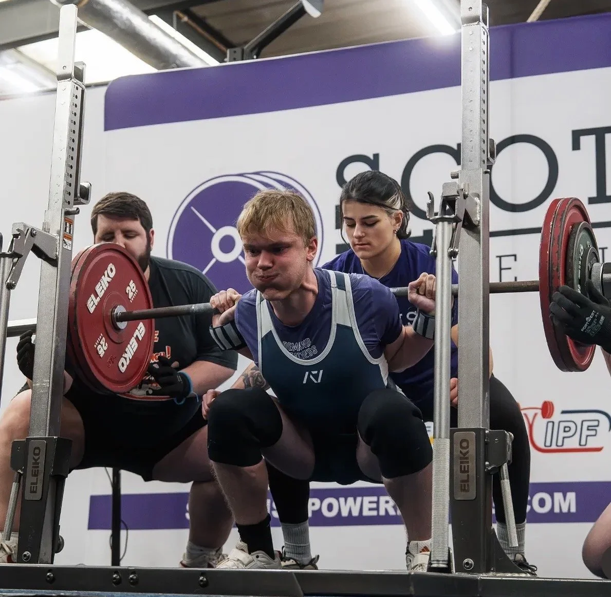 A powerlifter squatting with a loaded barbell, assisted by two spotters in a gym setting.