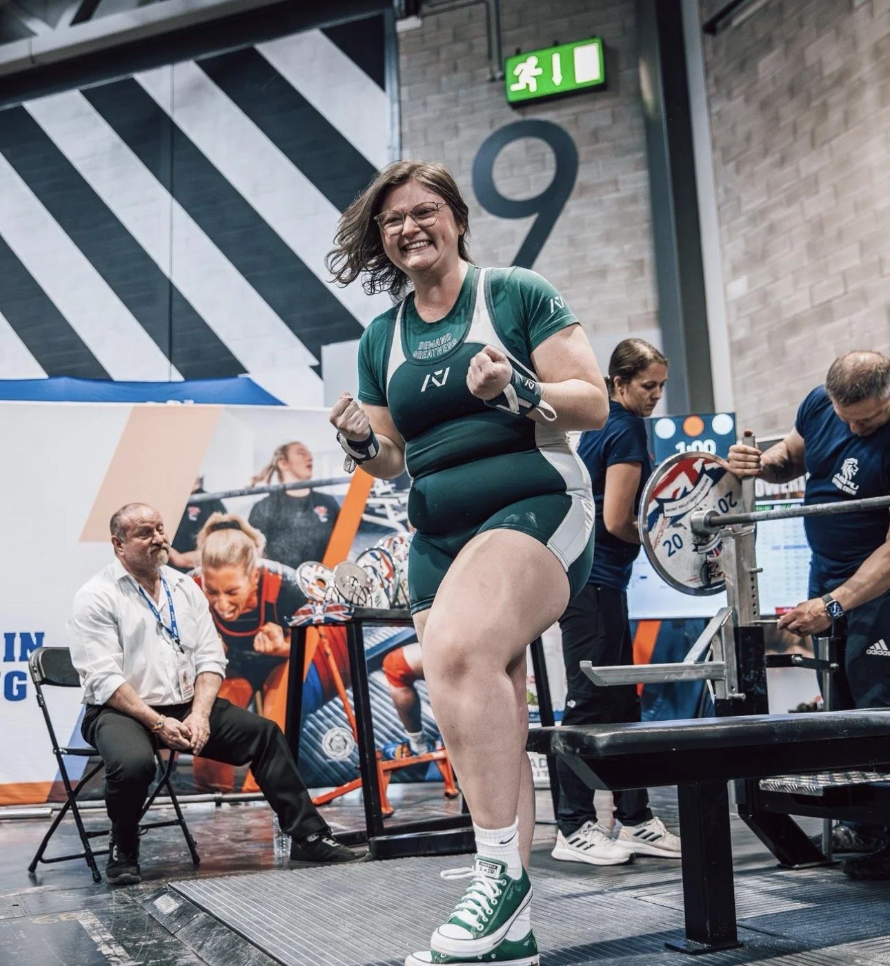 A woman celebrating after lifting weights at a powerlifting competition, with a big smile and clenched fists, wearing a green and black athletic outfit and sneakers, in a gym setting with other people and a chair nearby.
