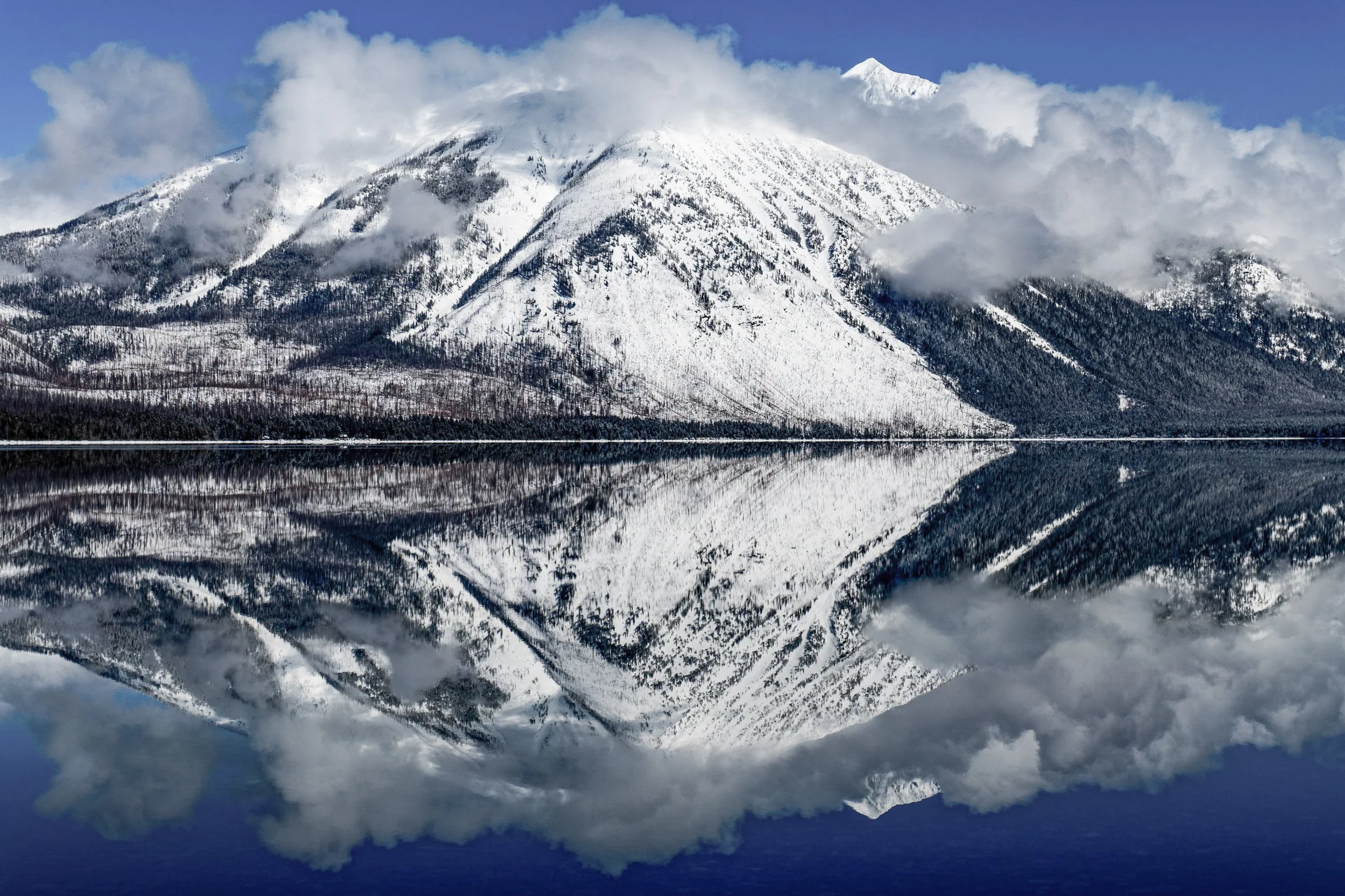 Snow-covered mountain with clouds around the peak, reflected in a calm body of water.