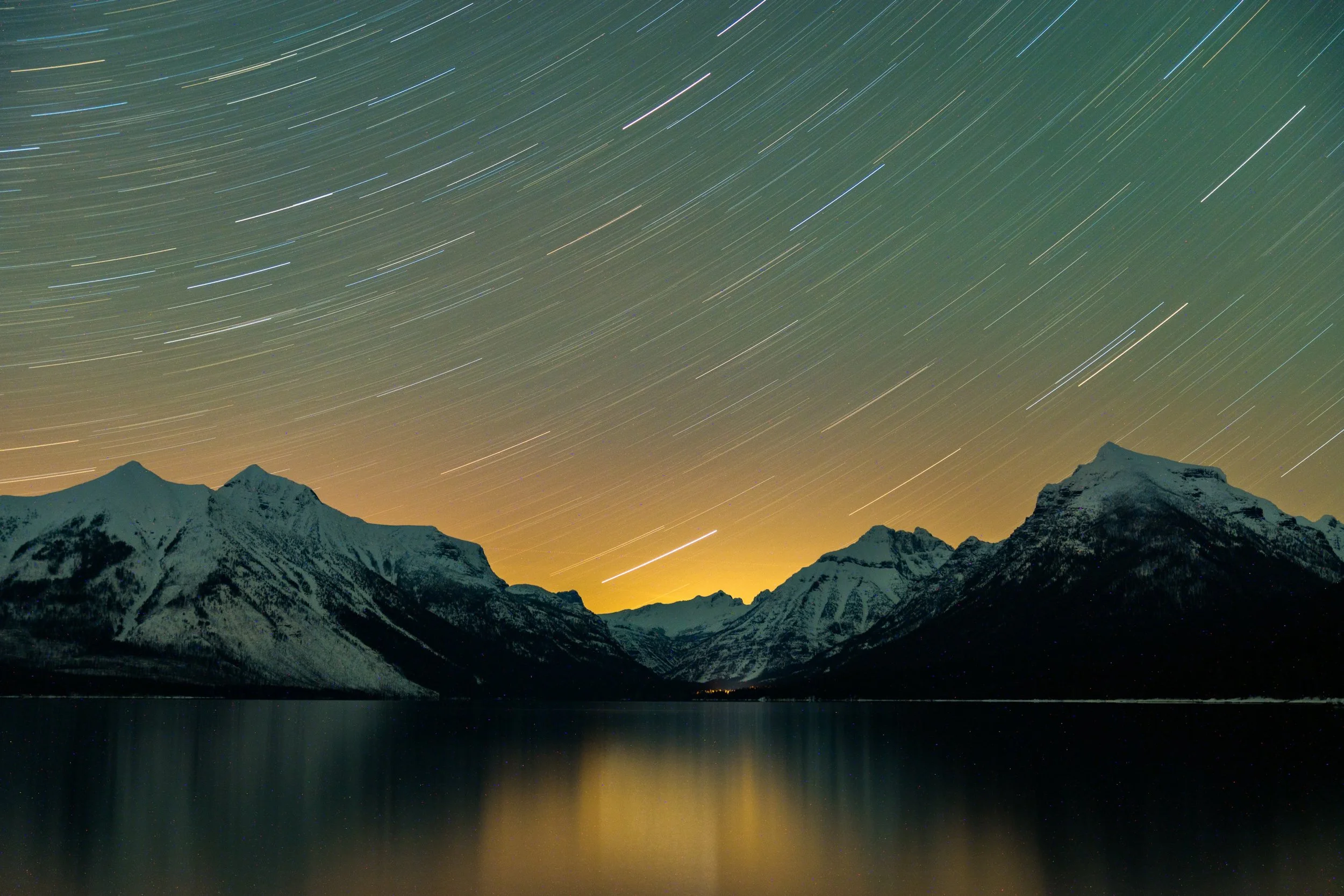 Nighttime mountain landscape with star trails in the sky and a body of water reflecting the mountains and star trails.