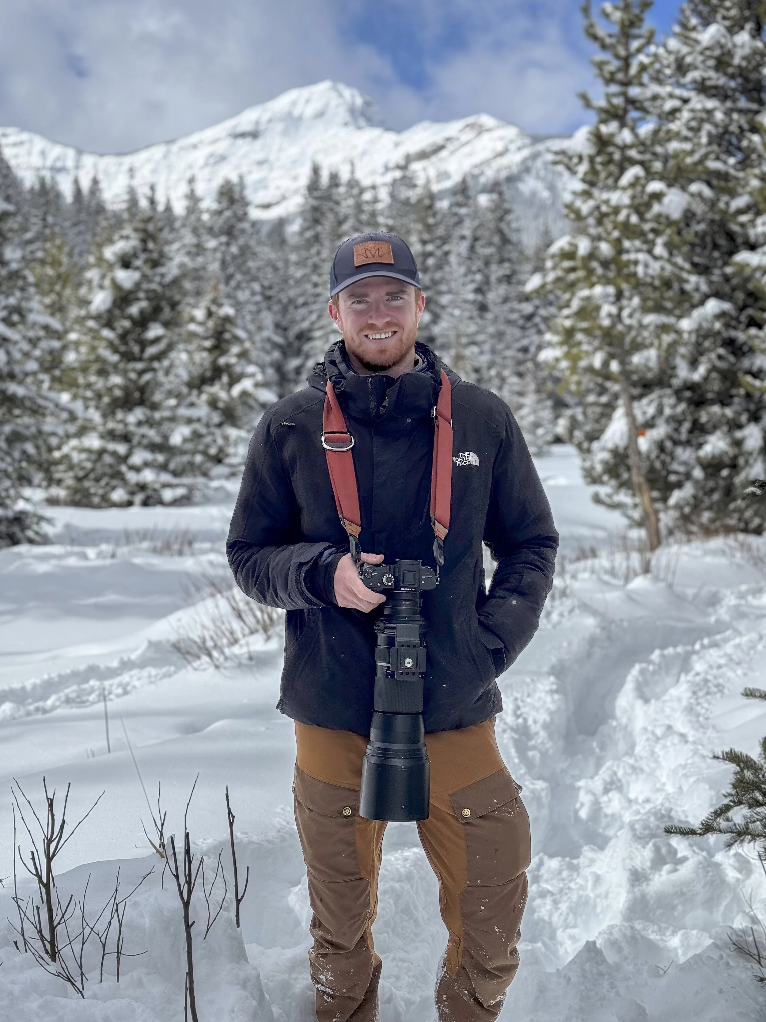 Man in winter outdoor clothing holding a camera, standing in snow-covered landscape with snowed trees and mountains in the background.