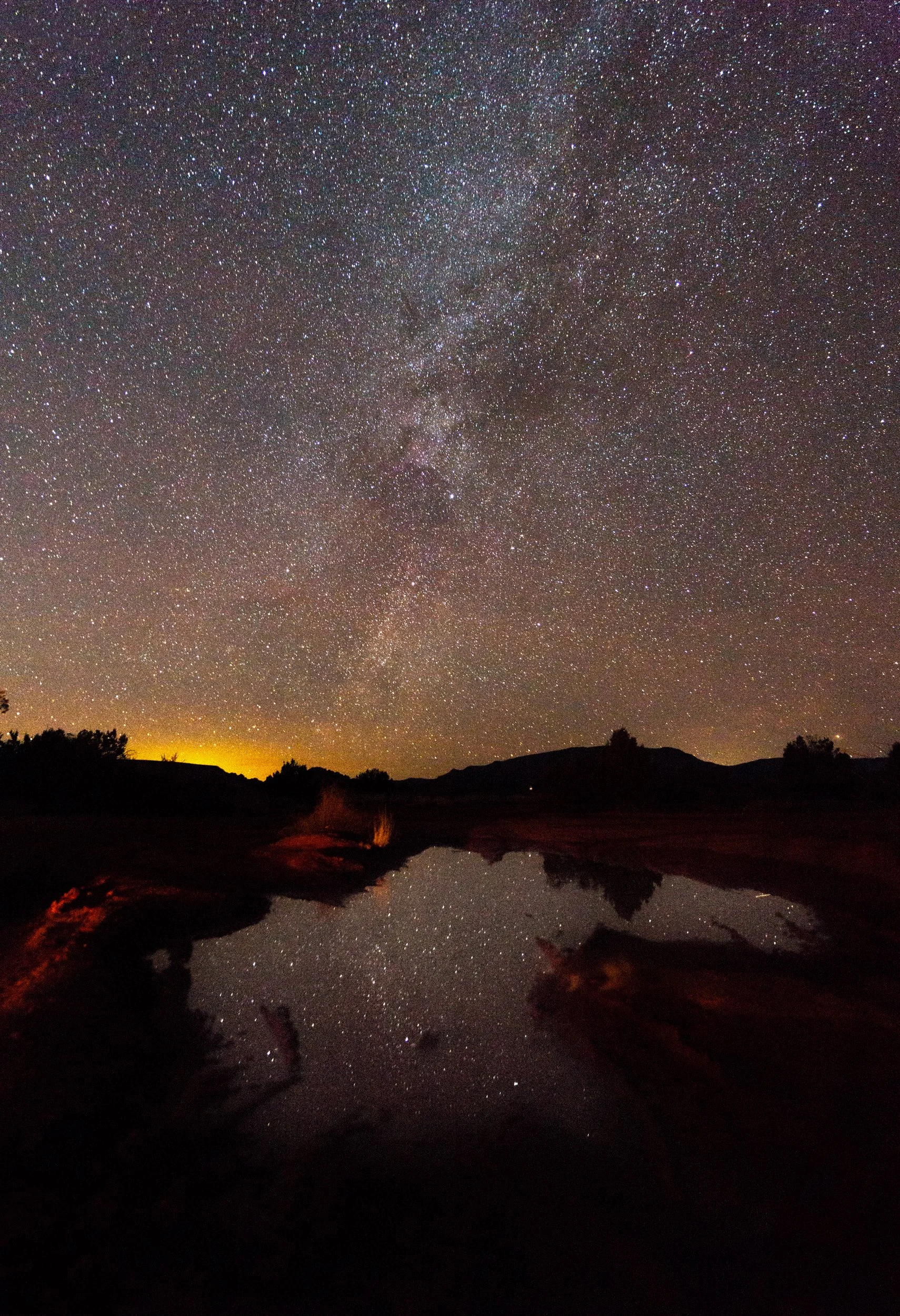 Night sky filled with countless stars and the Milky Way galaxy, reflected on a calm body of water, with silhouettes of hills and trees along the horizon.