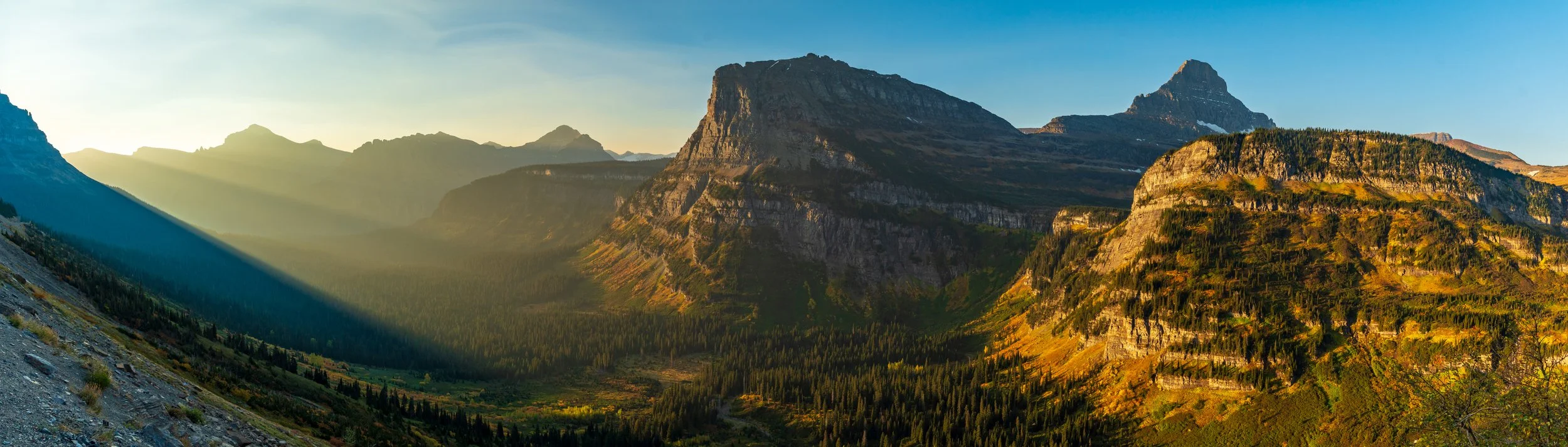 A panoramic view of a mountain valley with sunlit peaks, dense forest, and a green valley floor during sunset.